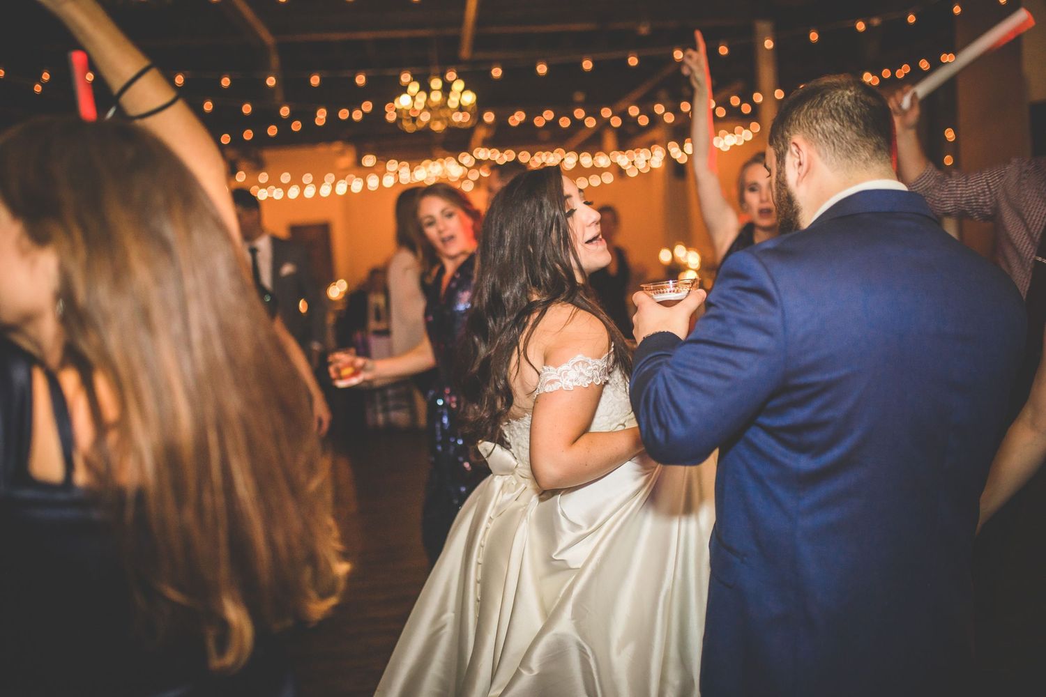 Wedding celebration with guests dancing under warm string lights and party atmosphere.