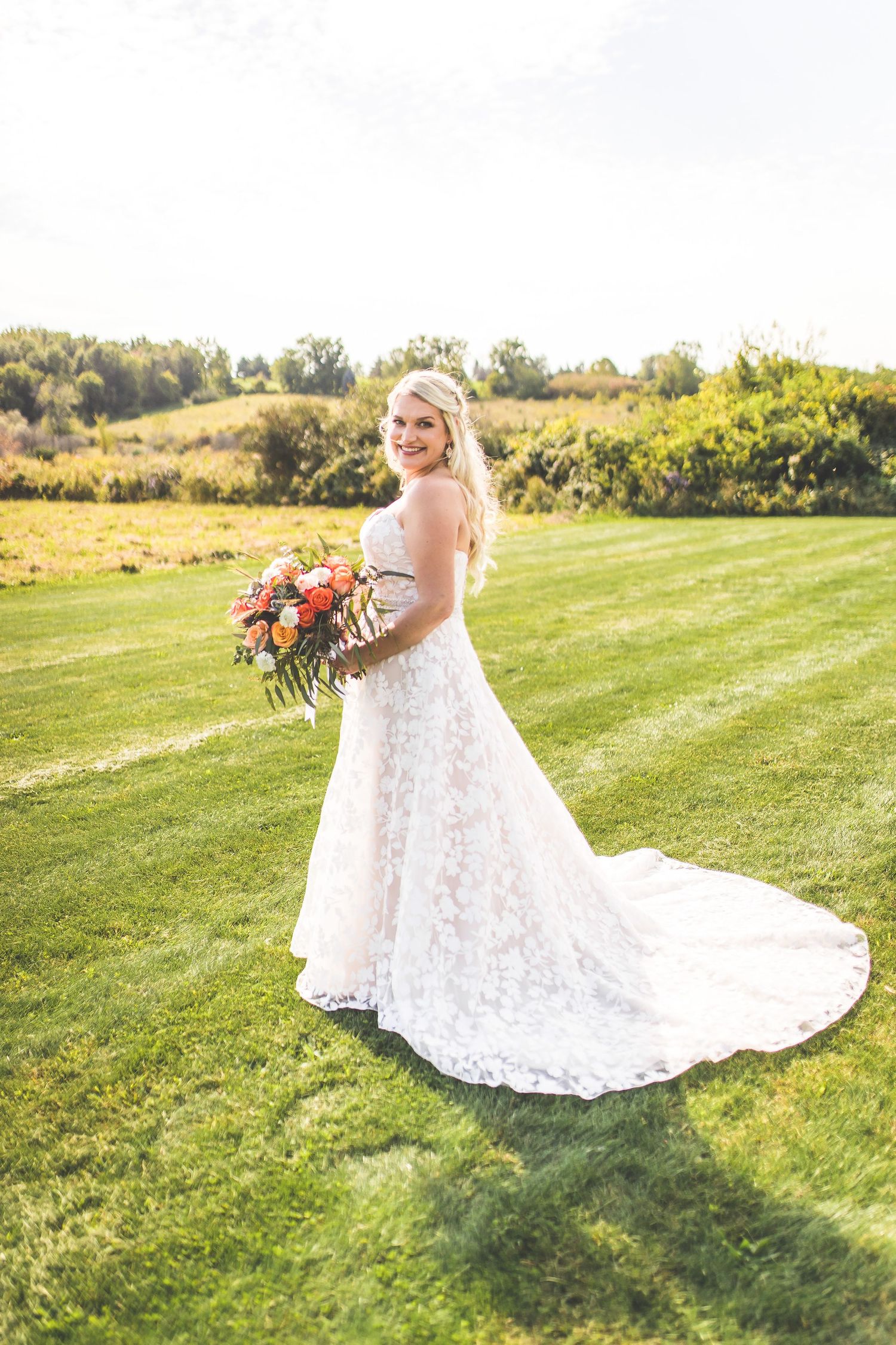 Bride in lace gown with train poses on sunlit grassy hillside.