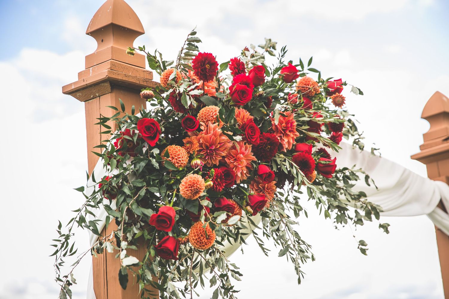 Wedding arch decorated with lush red and pink floral arrangements against blue sky.