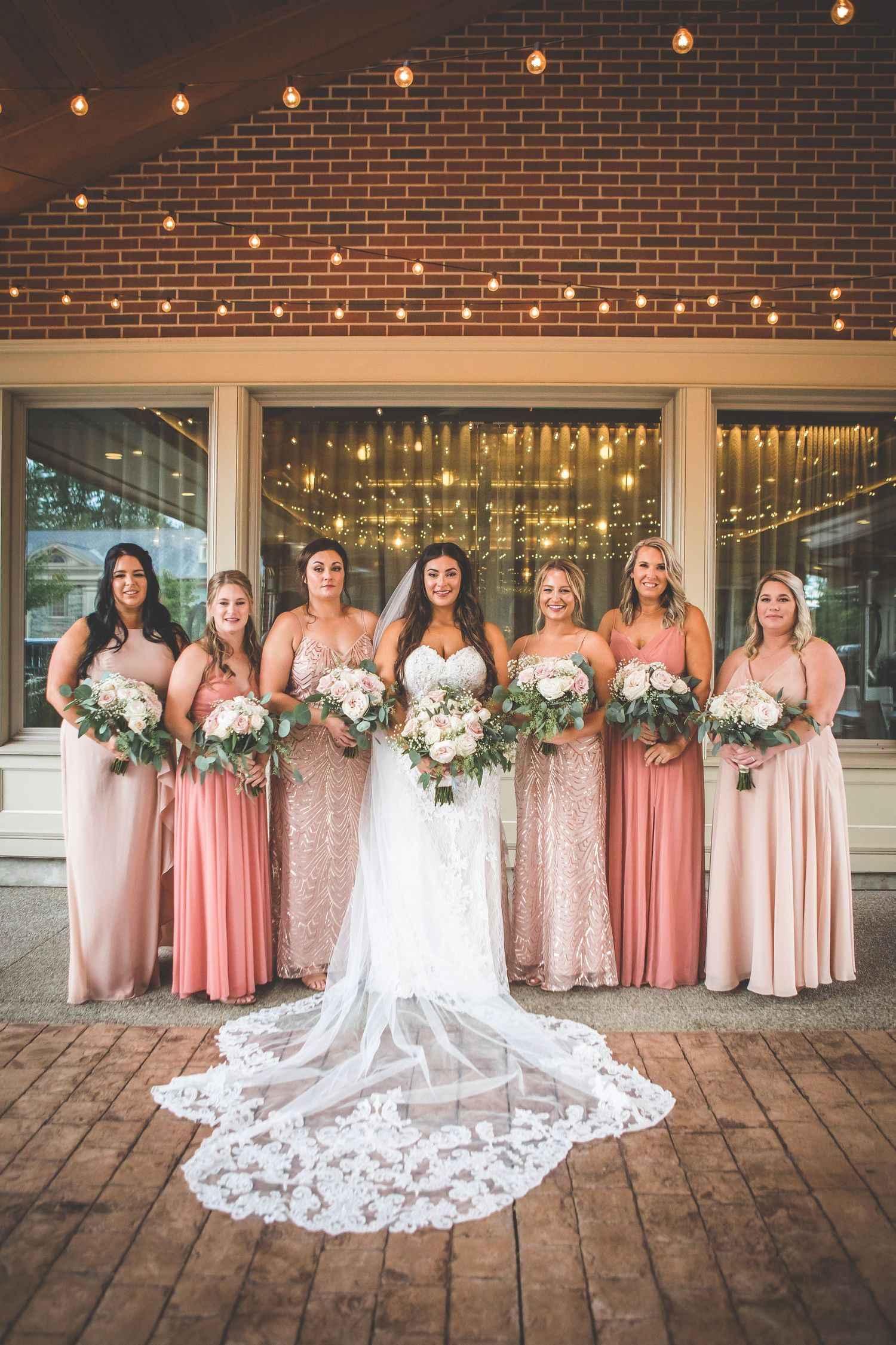 Bridal party in pink dresses poses under string lights on brick patio.