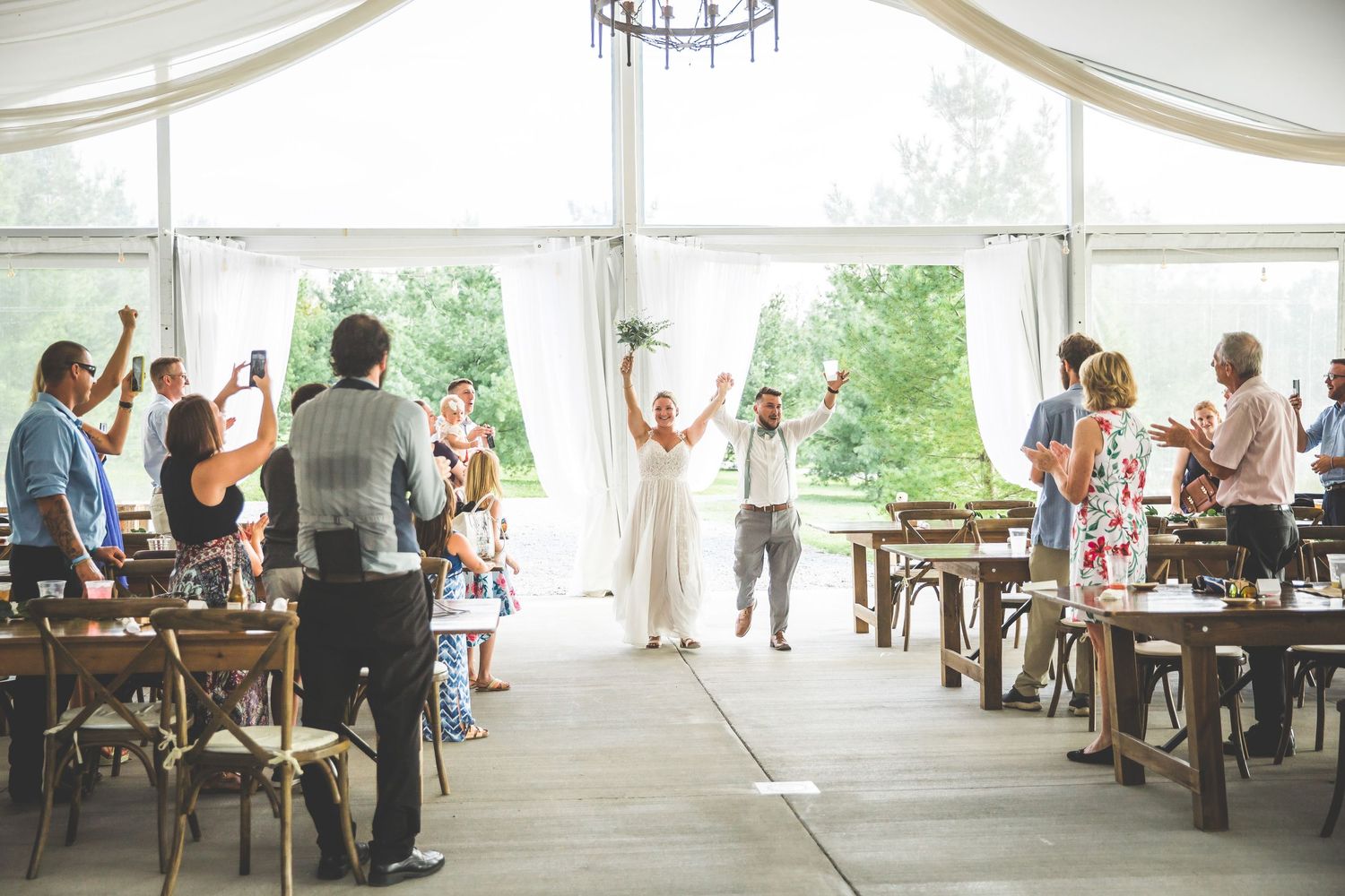 Elegant wedding reception setup inside a spacious white tent with rustic wooden tables and draping.
