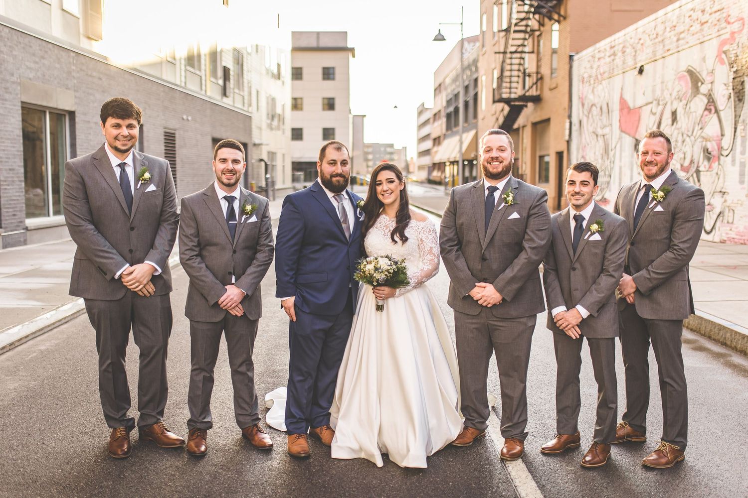 Wedding party with groomsmen in gray suits stand together on urban street.