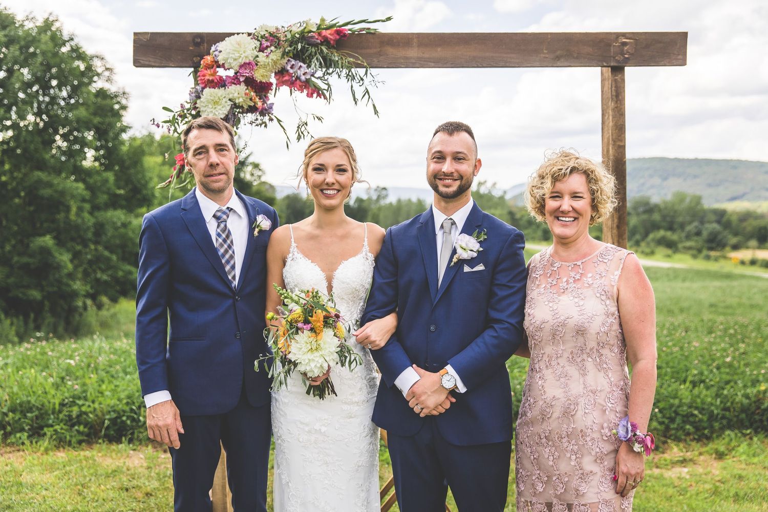 Wedding party members in navy suits and pink dresses gather under rustic wooden ceremony arch.