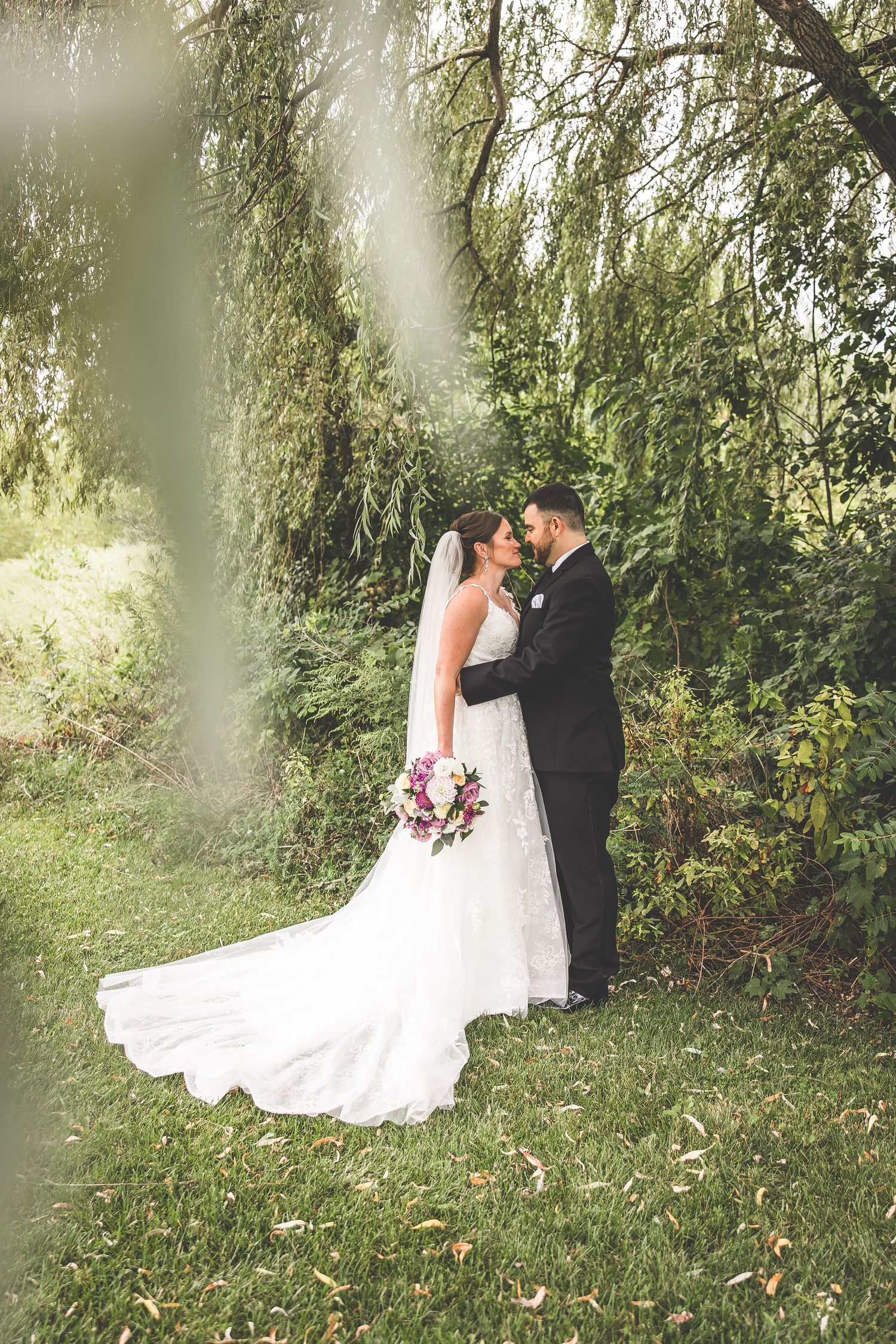 A romantic wedding couple embraces in a lush garden setting with willow trees creating a dreamy backdrop.