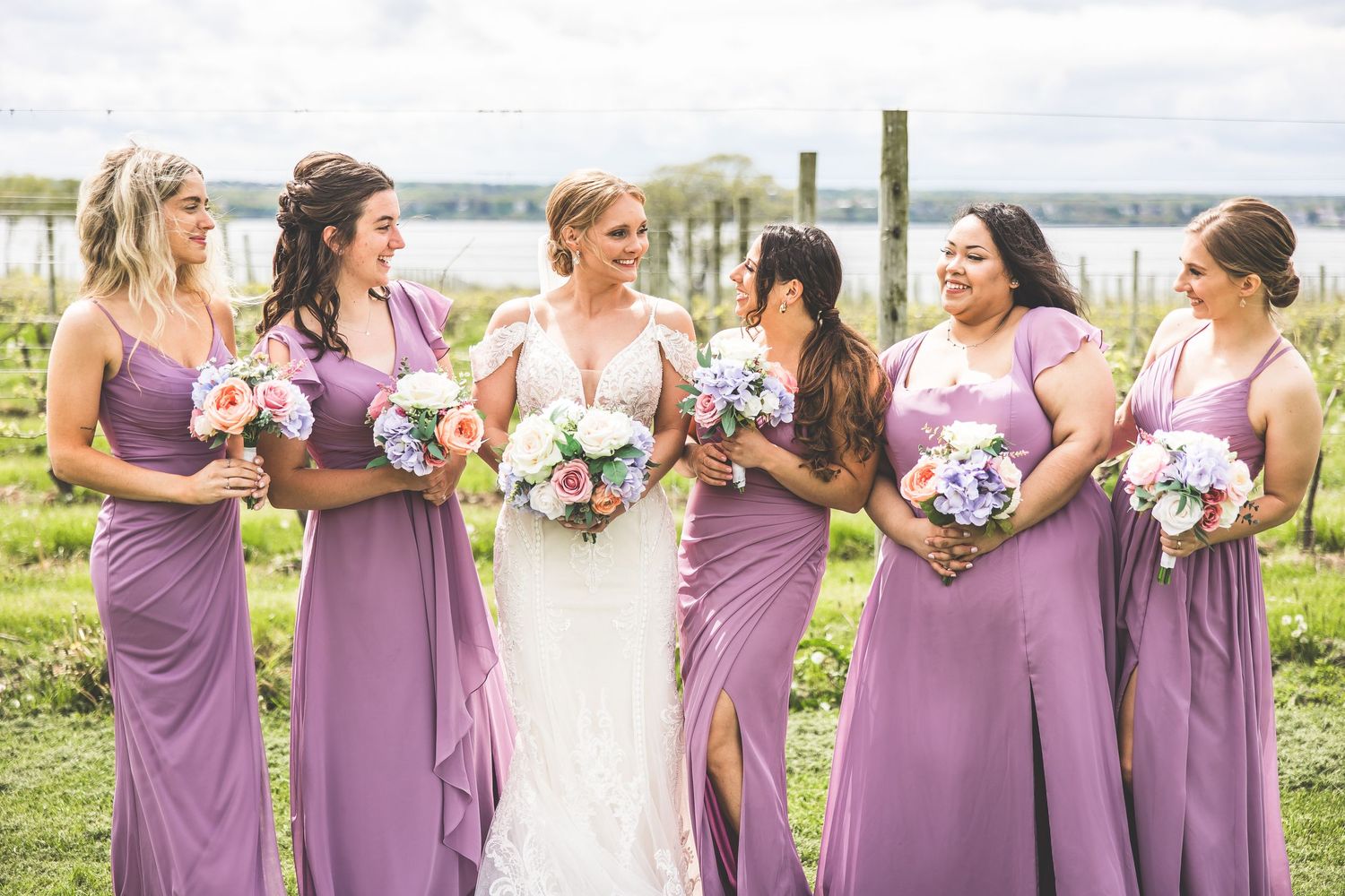 Wedding party in purple dresses poses with bouquets by waterfront.