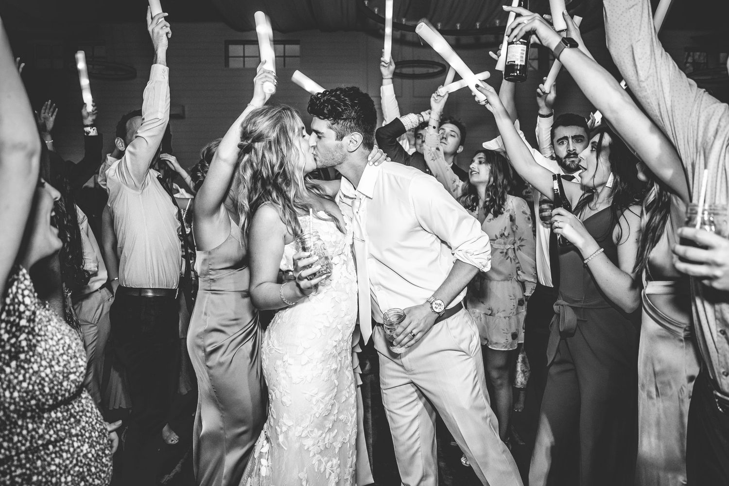 Wedding reception guests dancing and celebrating on a crowded dance floor with raised hands and glowing light sticks.