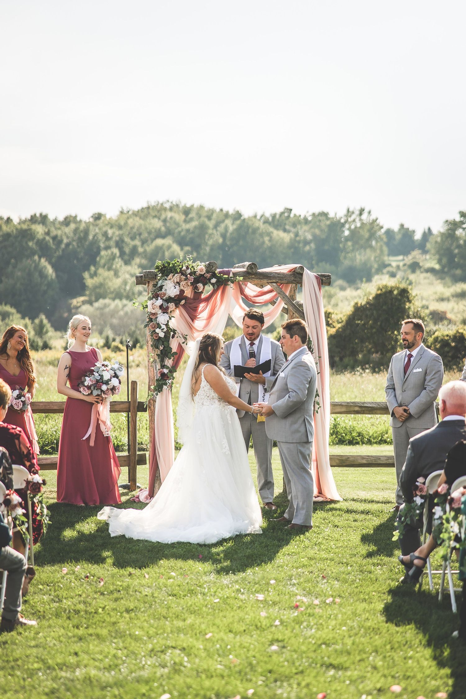 Sunlit outdoor wedding ceremony with guests watching couple exchange vows under floral-decorated arch.