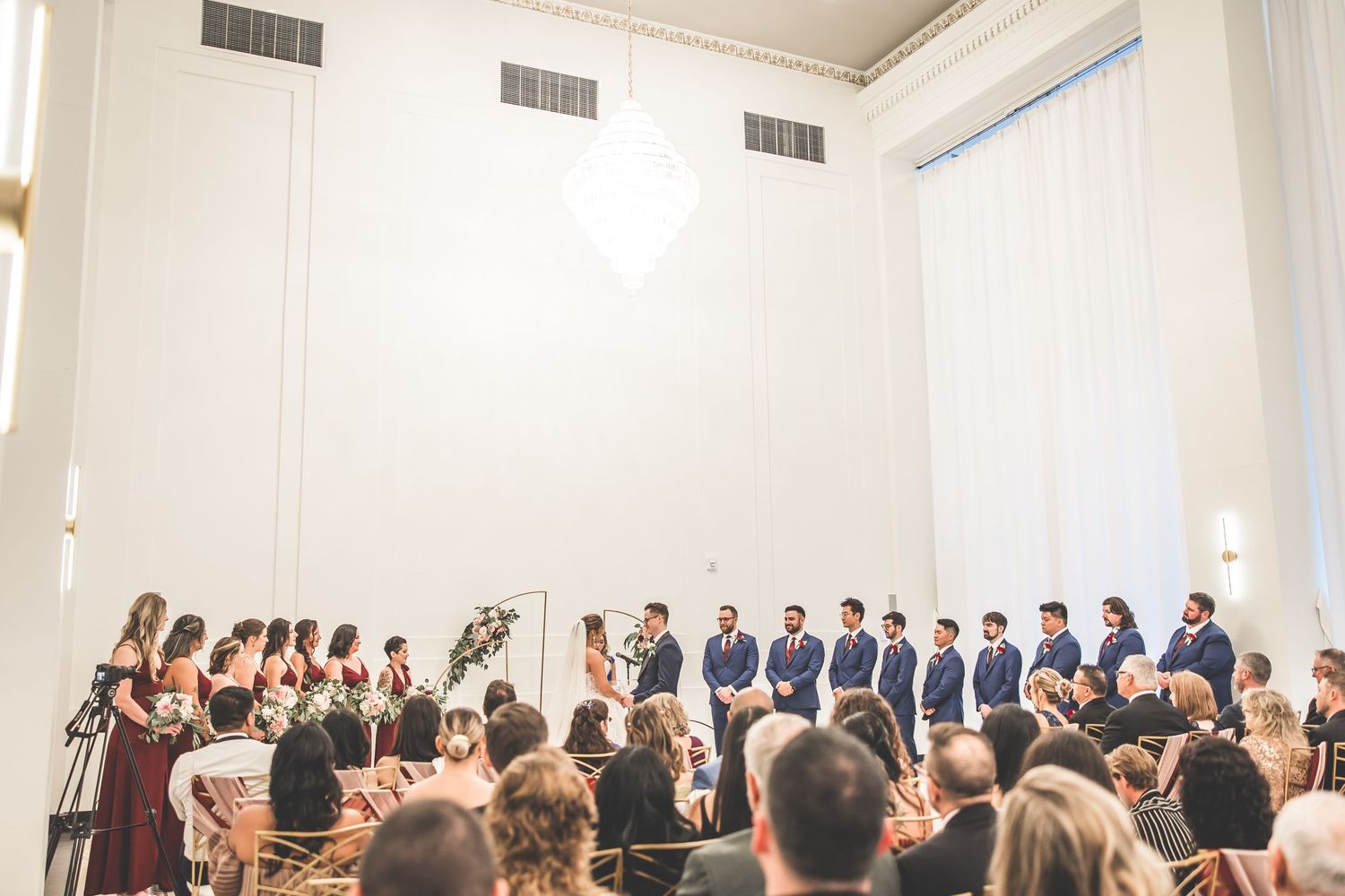Panoramic view of wedding guests seated during ceremony in bright white venue.