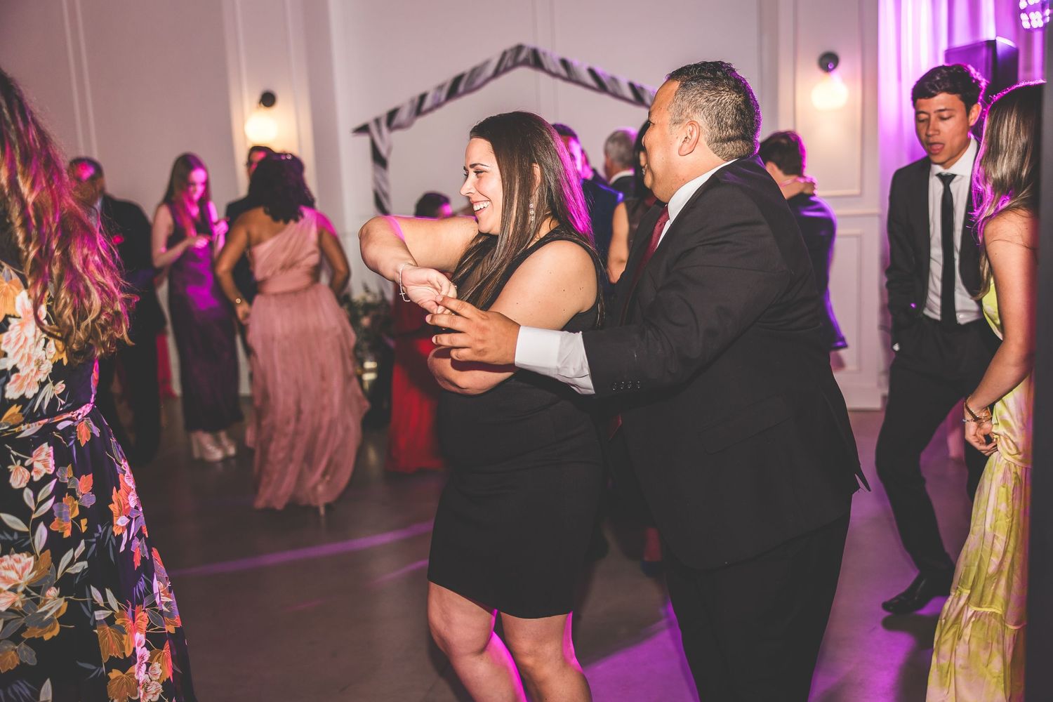 Couples enjoying dances at a wedding reception with purple mood lighting and white drapes.