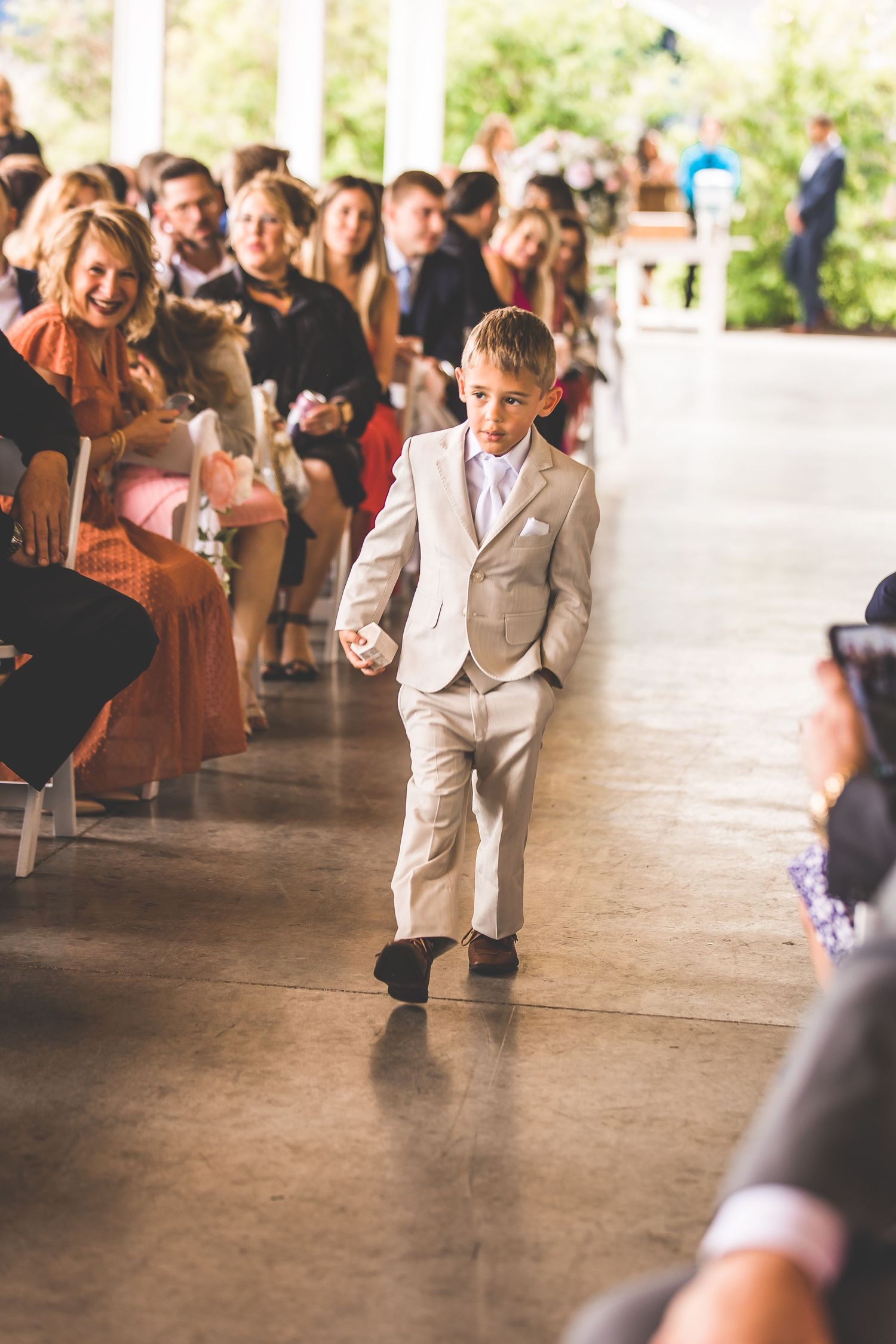 Young ring bearer in light colored suit walks down aisle during wedding ceremony.