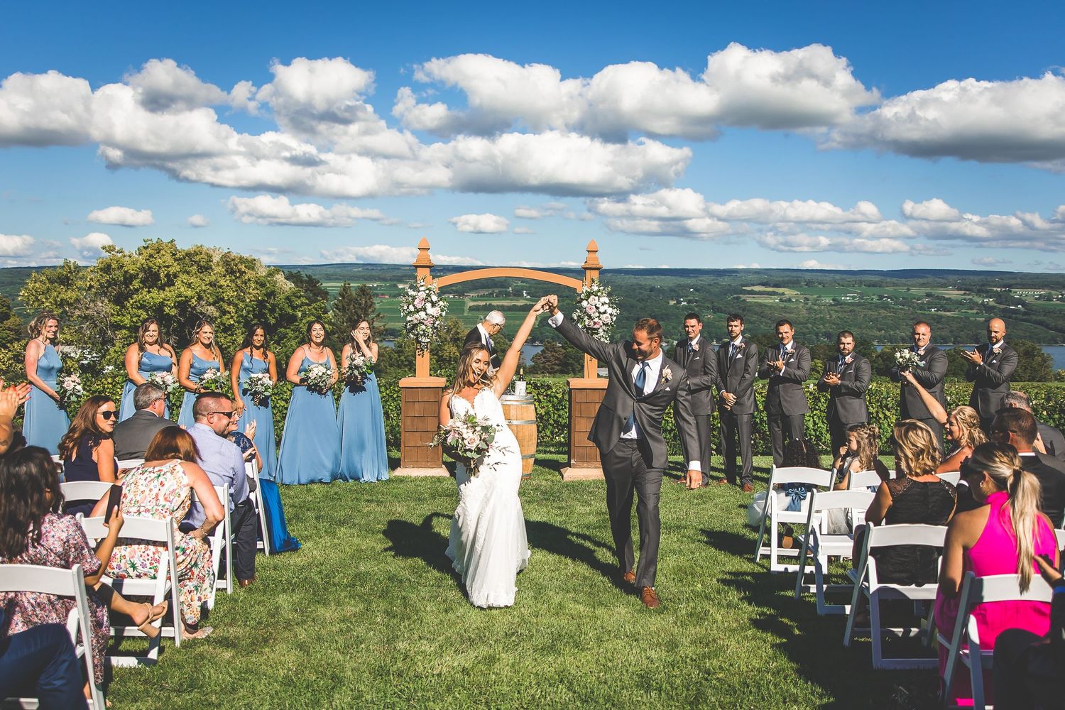 Panoramic view of outdoor wedding ceremony setup with white chairs and guests against rolling hills landscape.