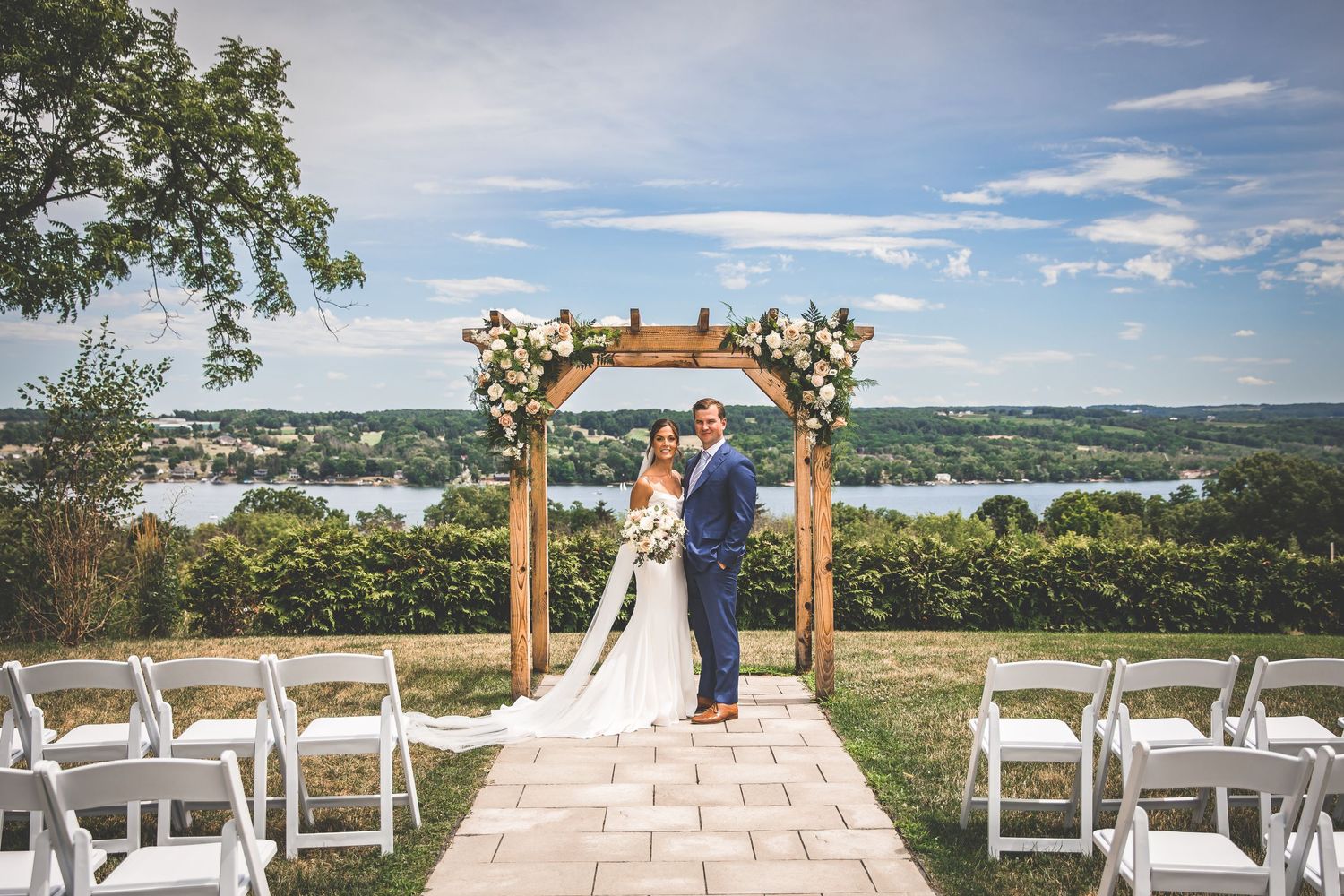 Outdoor wedding ceremony setup with wooden arch and white chairs overlooking a scenic lake view.