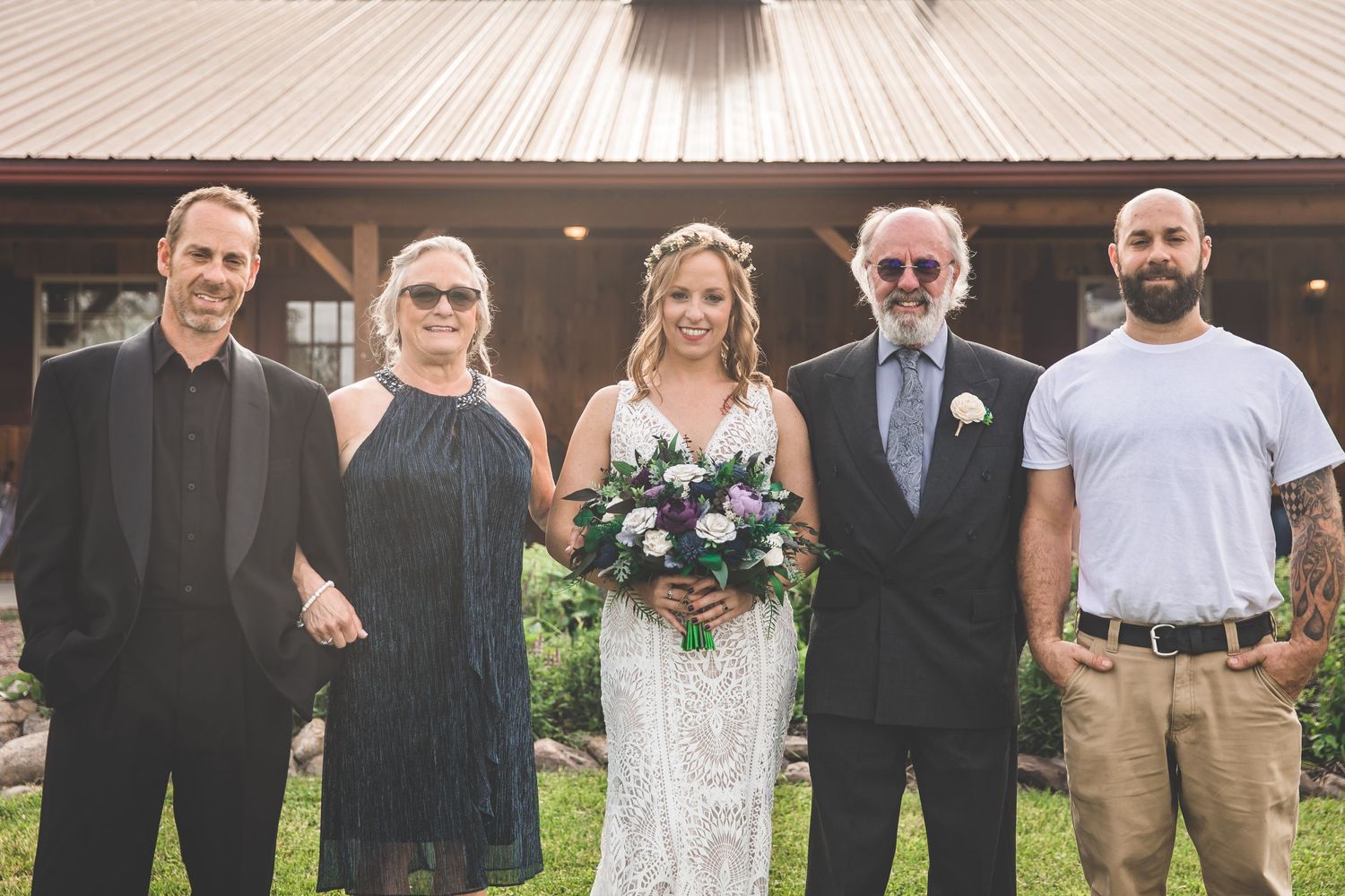 Wedding party group photo taken outdoors with rustic barn in background.