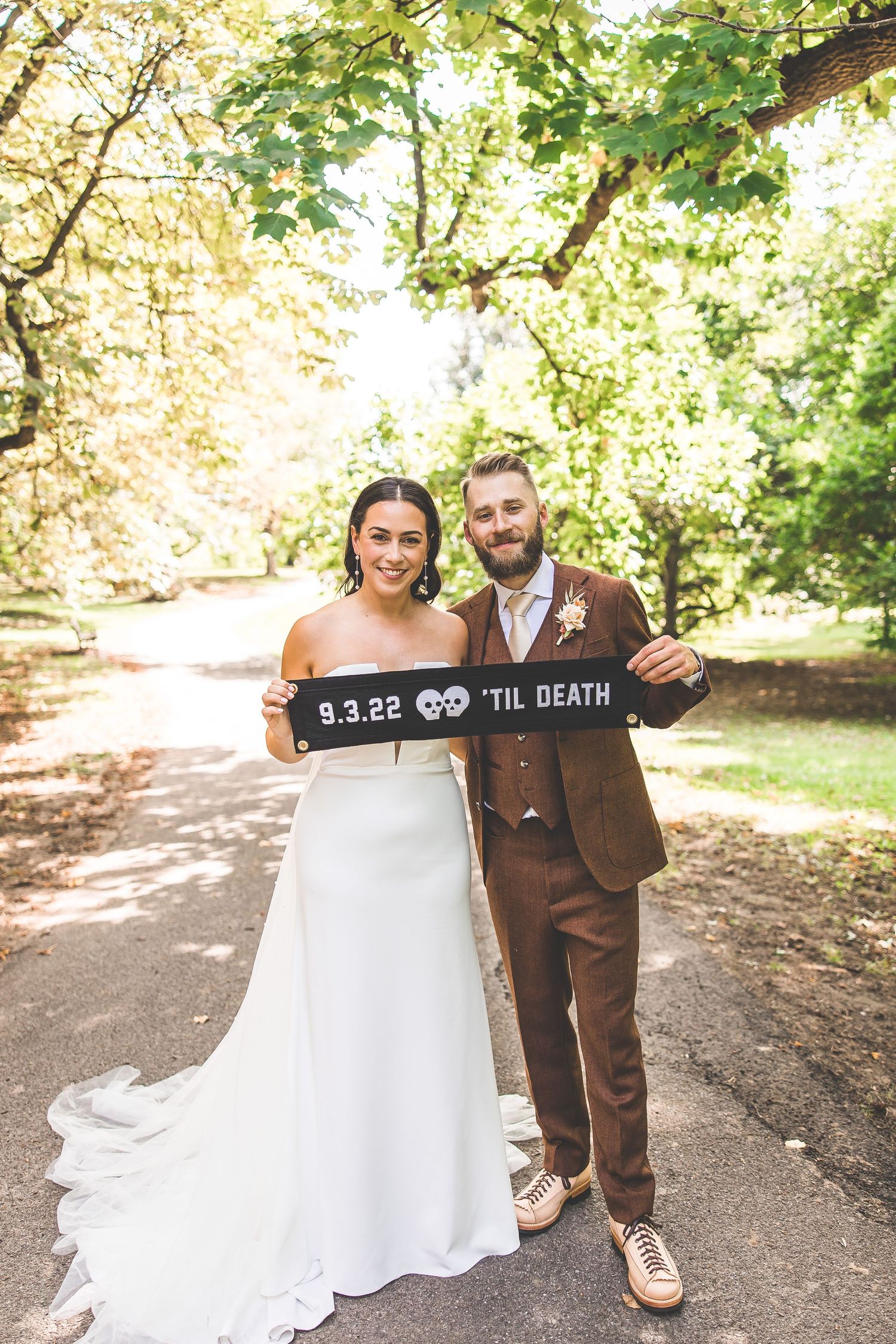 Newlyweds hold 'Til Death' sign on garden path in wedding attire.