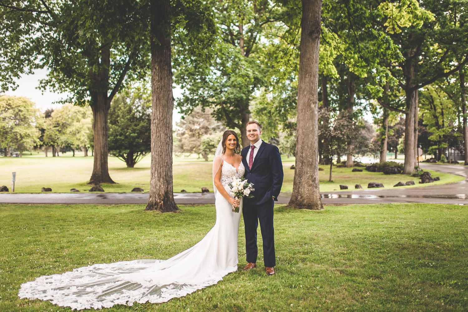 Outdoor wedding portraits taken under tall trees in a scenic park setting.