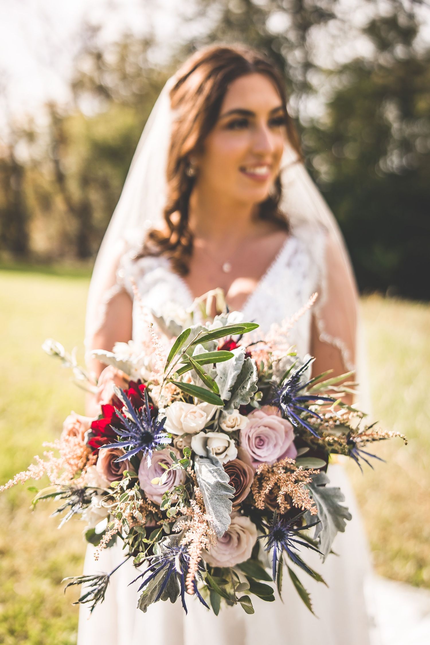 Close-up of bride's rustic bouquet featuring roses, thistles, and greenery in natural sunlight.