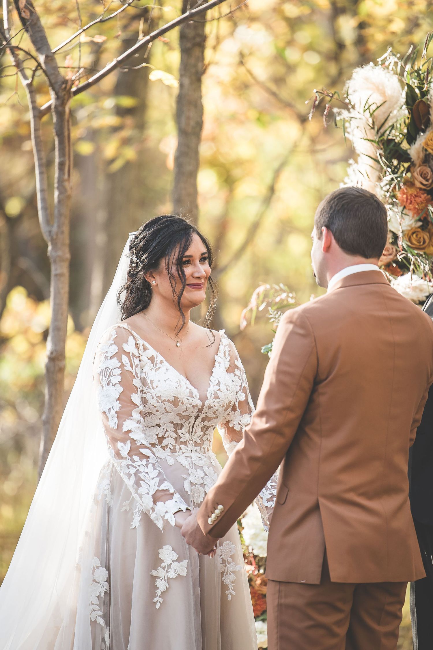 Elegant outdoor wedding ceremony with bride in lace dress and groom in brown suit among autumn trees.
