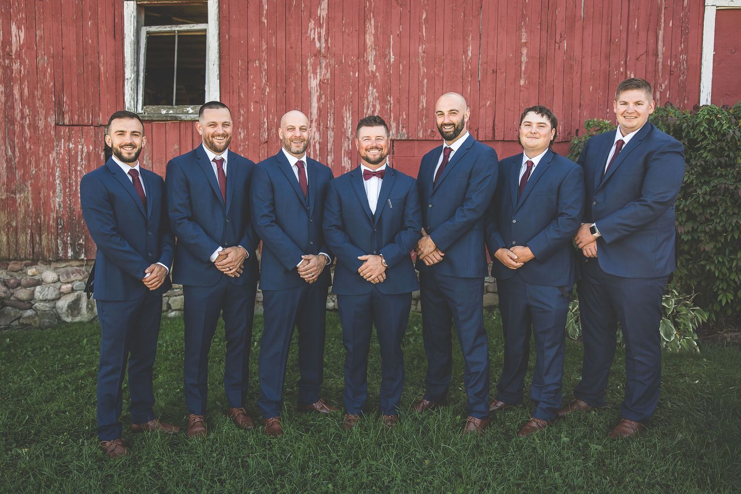 Groomsmen in navy suits line up against weathered red barn wall for wedding photo.