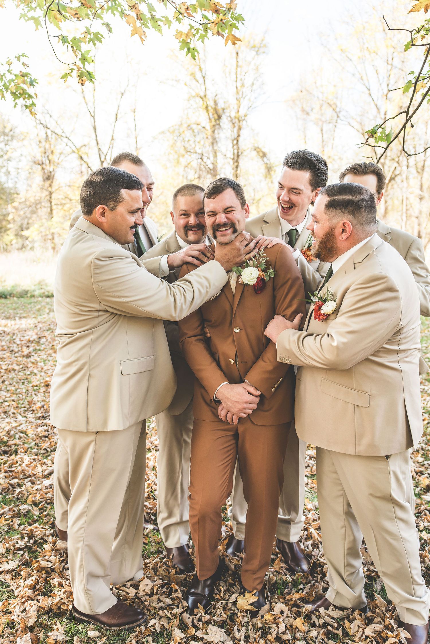 Groomsmen in beige suits playfully gather around the groom in brown during outdoor wedding photos.