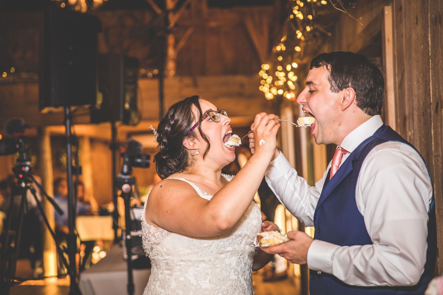 Wedding cake feeding tradition captured at rustic barn reception with string lights.