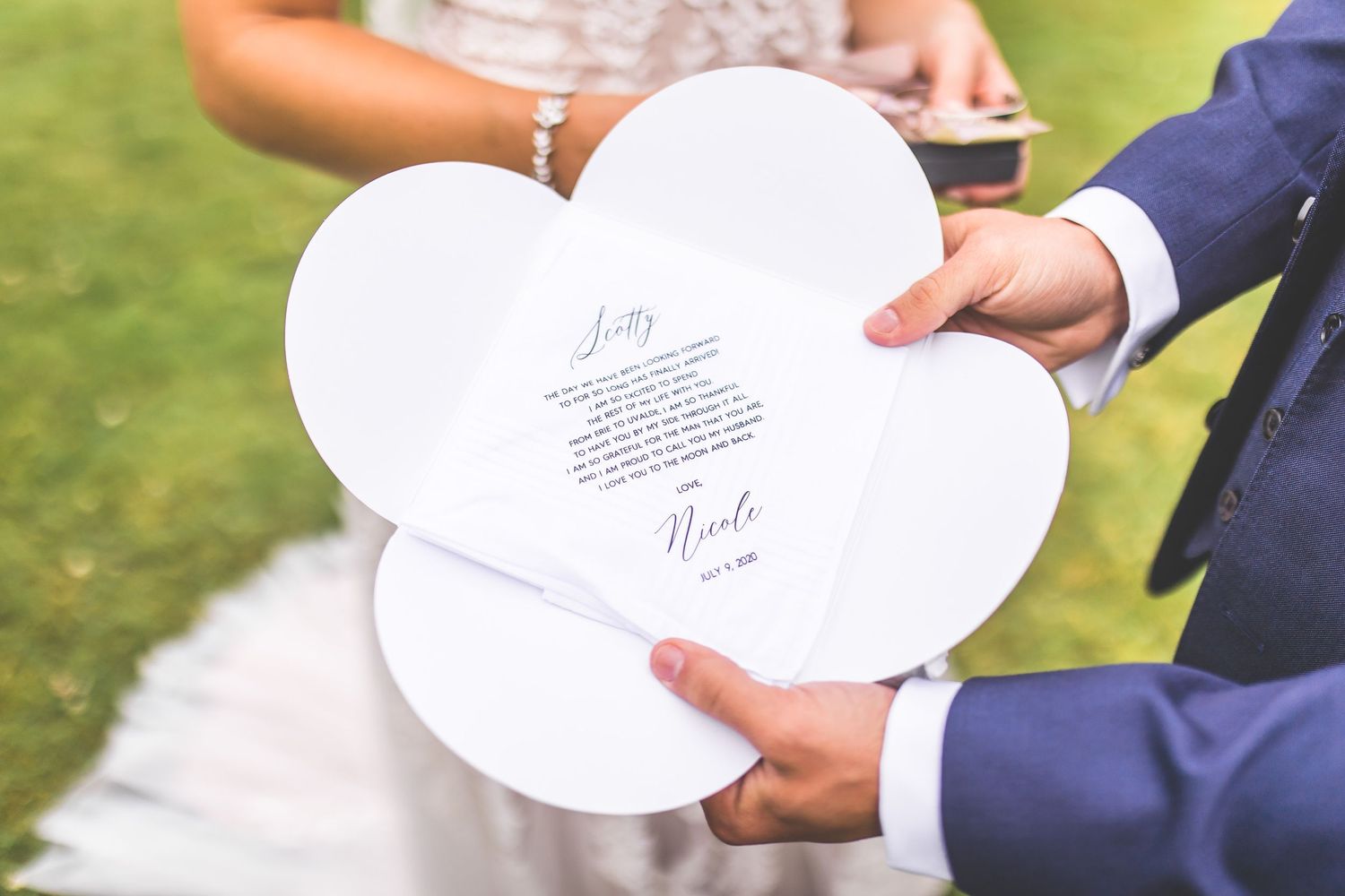 A white heart-shaped wedding vow booklet held between two people outdoors.