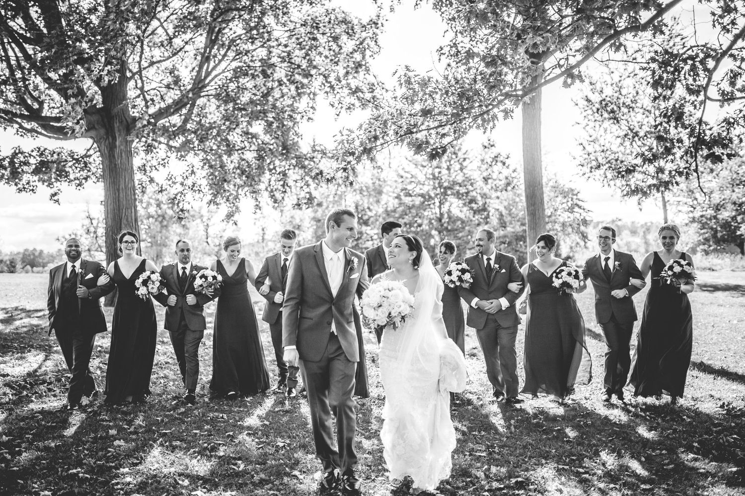 Large wedding party gathered outdoors under trees for formal group photo.