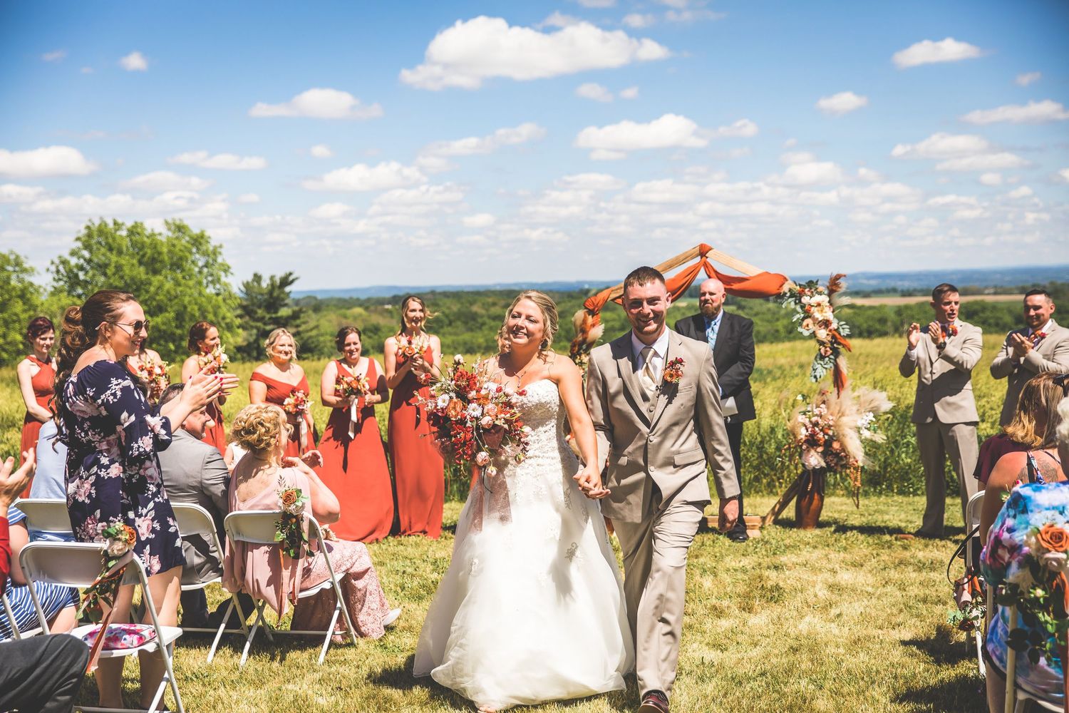 Panoramic view of outdoor wedding ceremony with guests seated on hillside under blue sky.