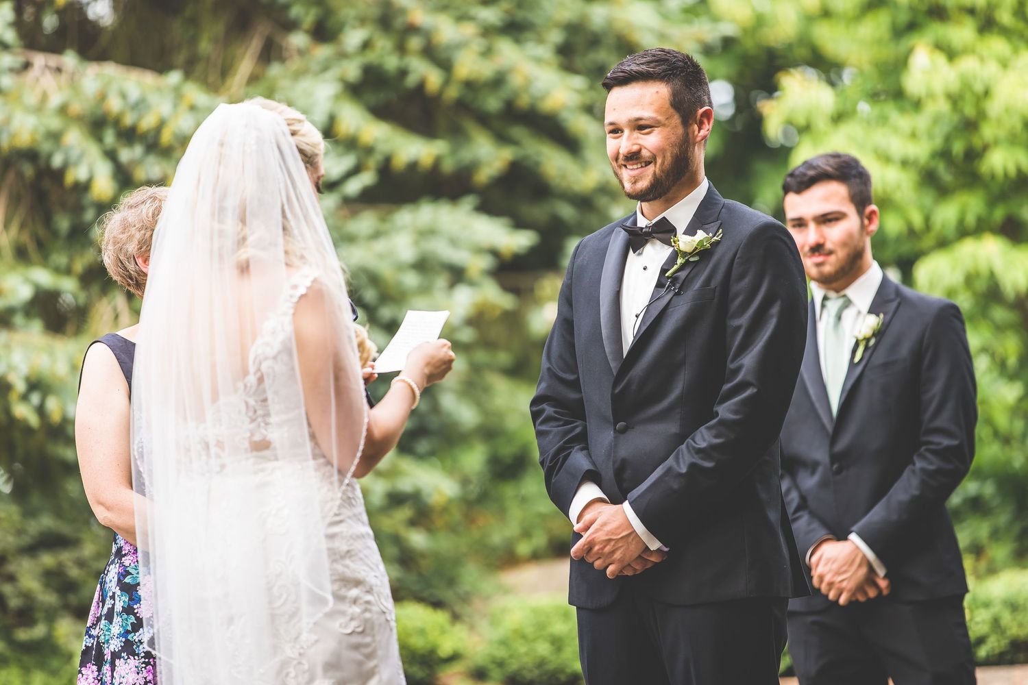 Wedding ceremony exchange of rings in a garden setting with evergreen trees and purple flowers in background.