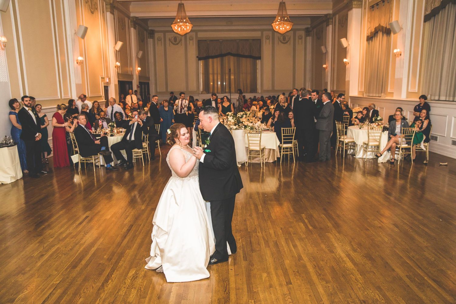 Couples dance together in an elegant ballroom with hardwood floors and ornate lighting during a wedding reception.