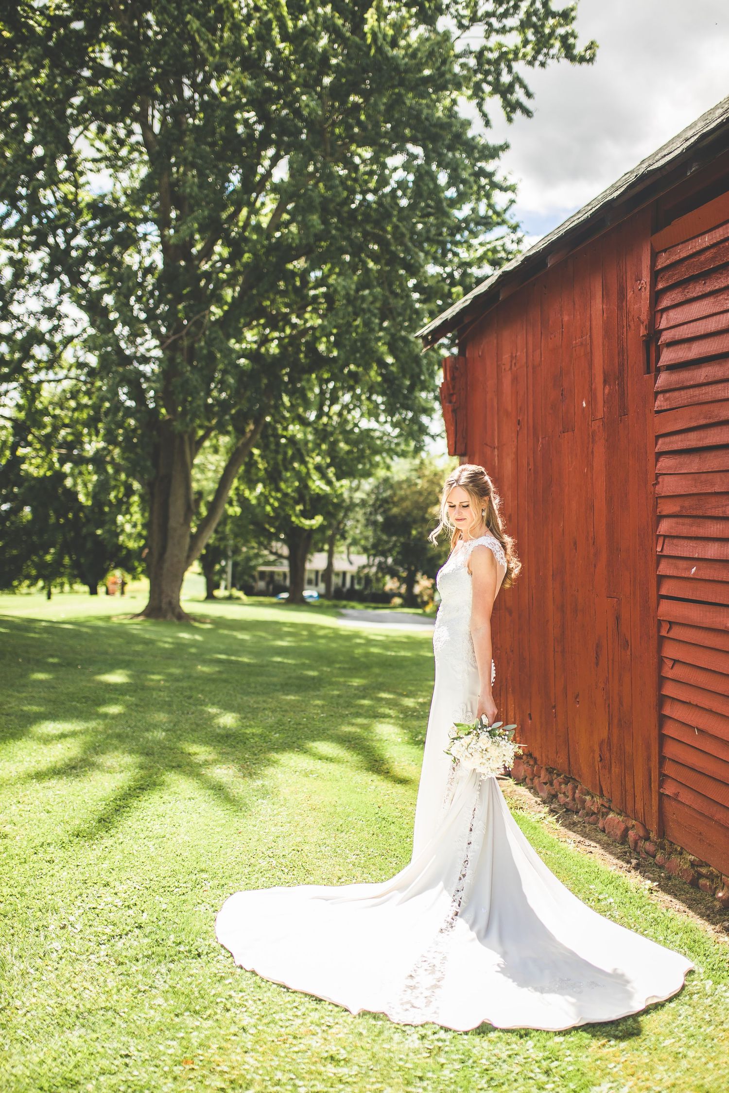 White wedding dress flowing behind bride against rustic red barn in sunlit garden.