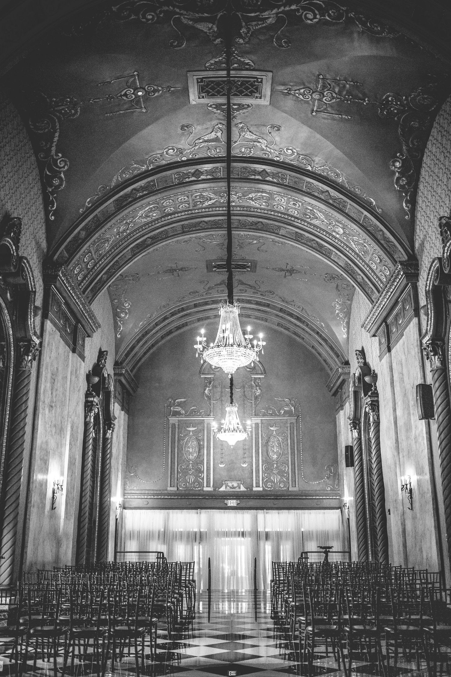 Dramatic black and white photo of ornate church interior with chandeliers and rows of chairs.