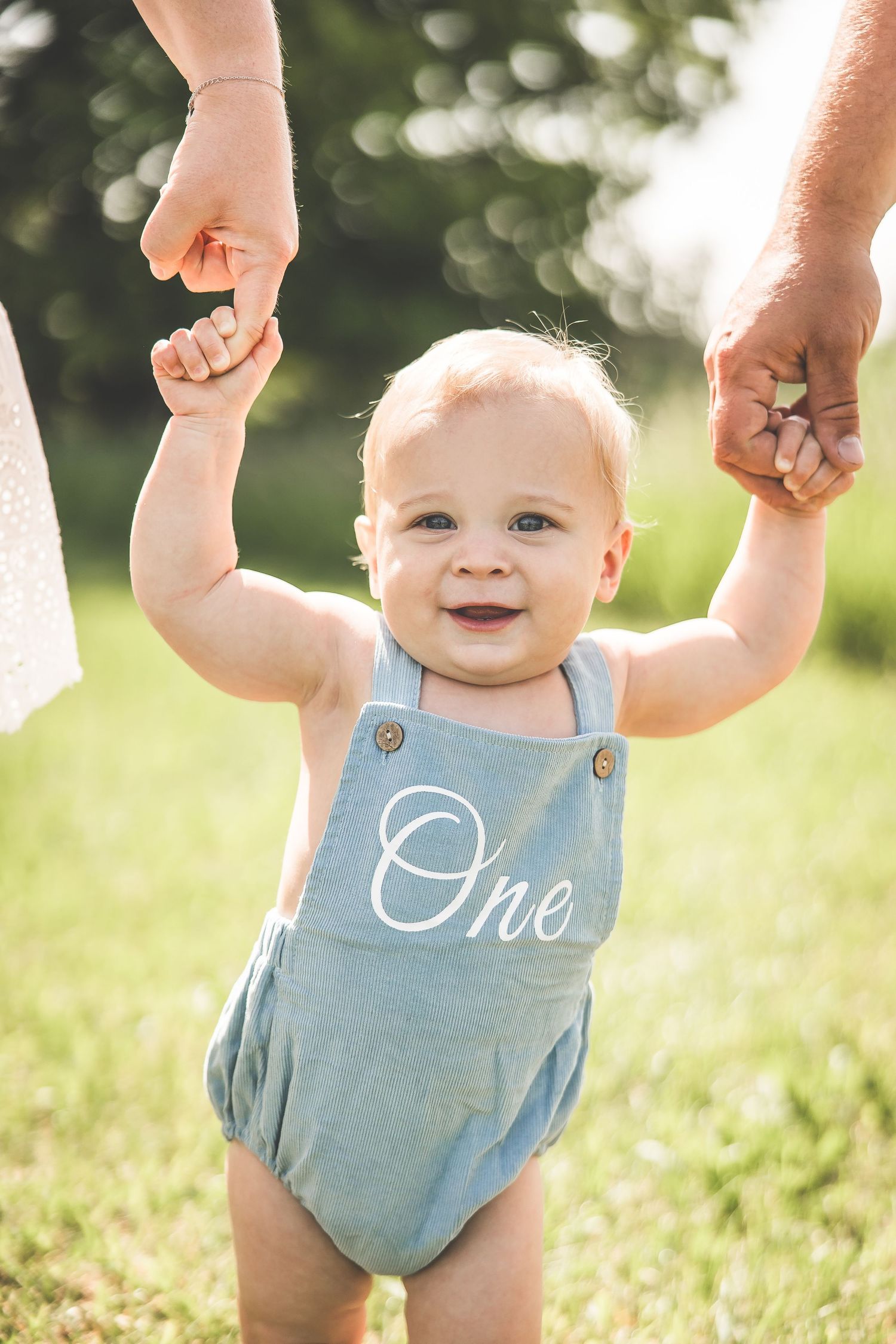 Baby in blue romper with 'One' text holding parents' hands while standing outdoors.