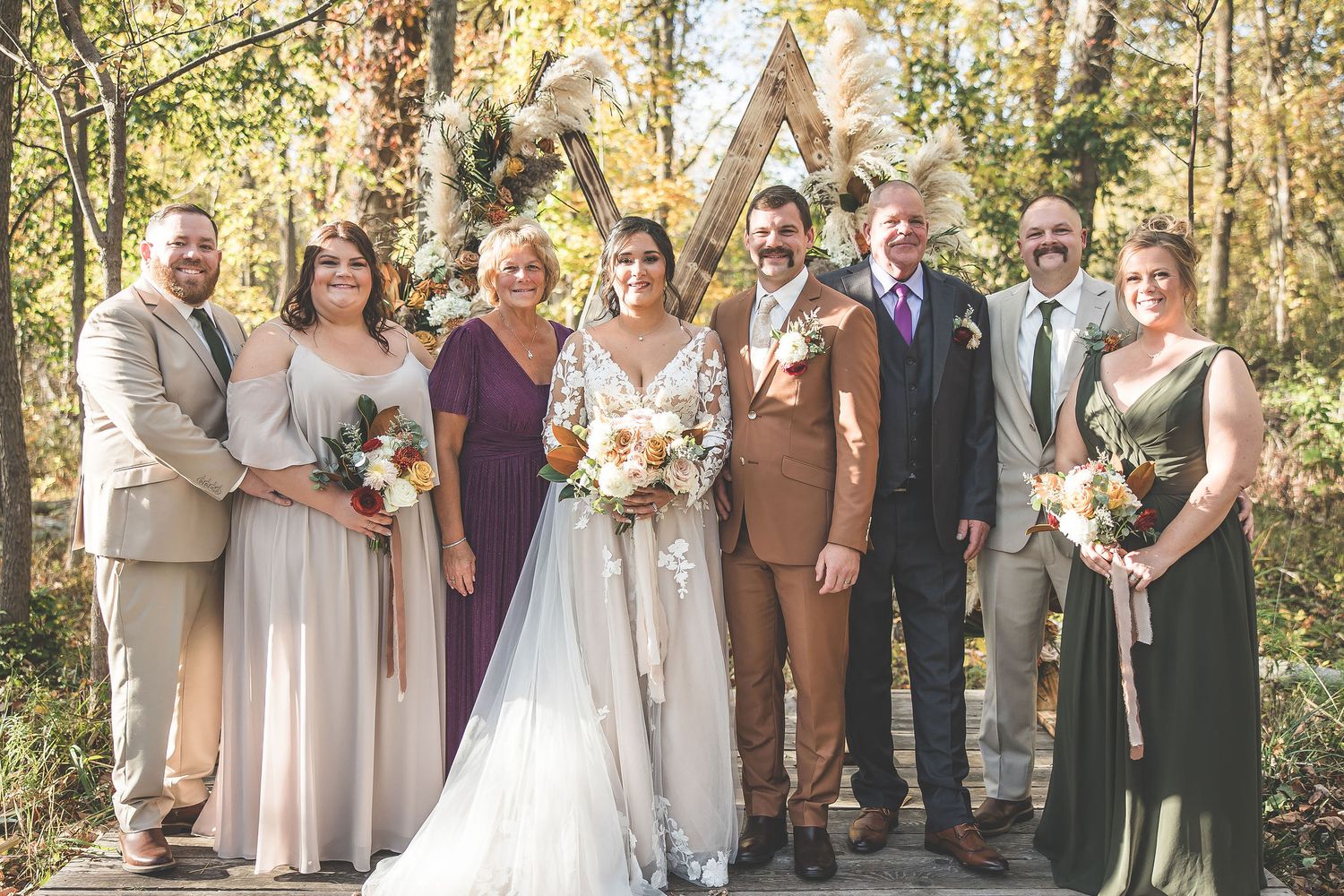 Wedding party stands together in a wooded area with fall foliage and sunlight filtering through trees.