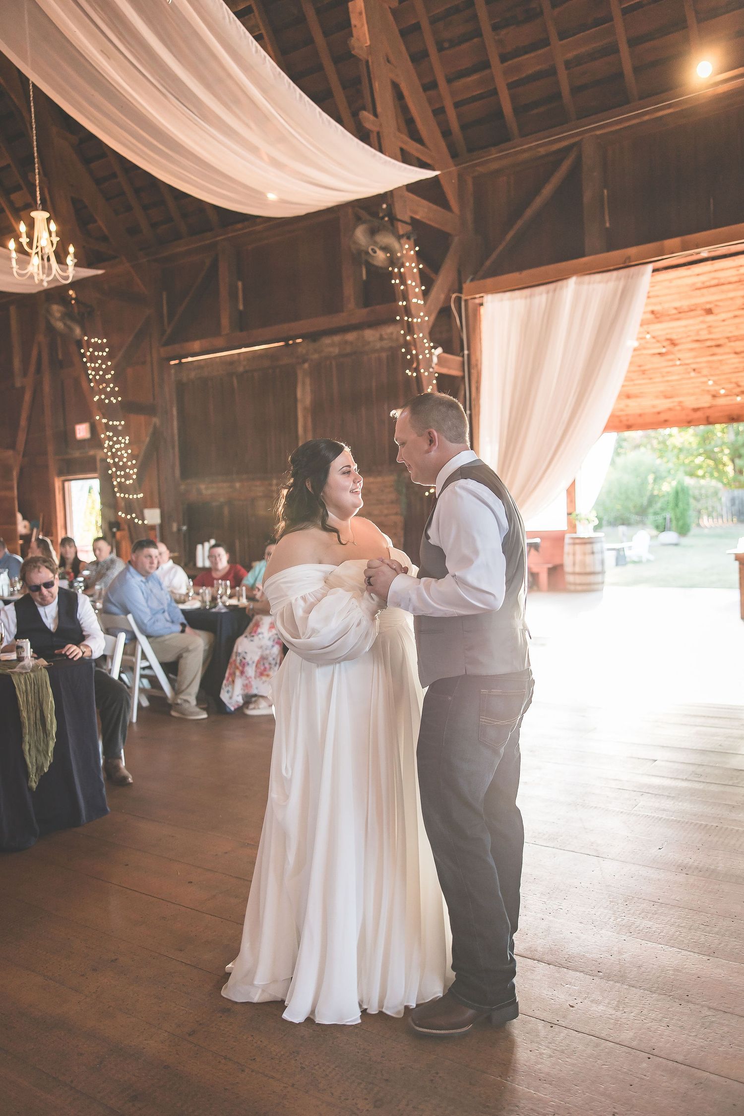 Wedding guests dance in a rustic barn venue decorated with flowing white curtains and string lights.