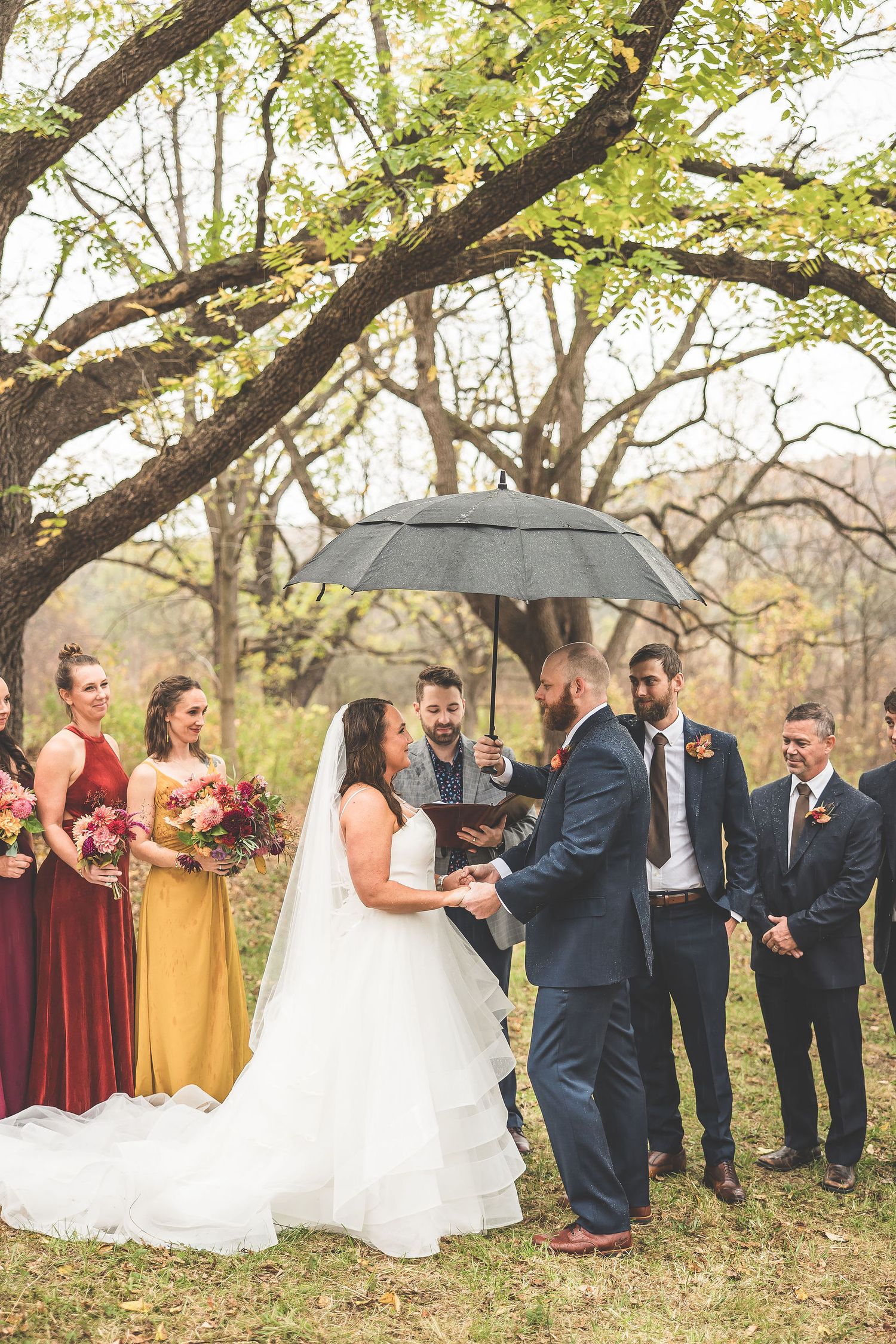 Wedding ceremony under autumn trees with black umbrella and burgundy bridesmaid dresses.