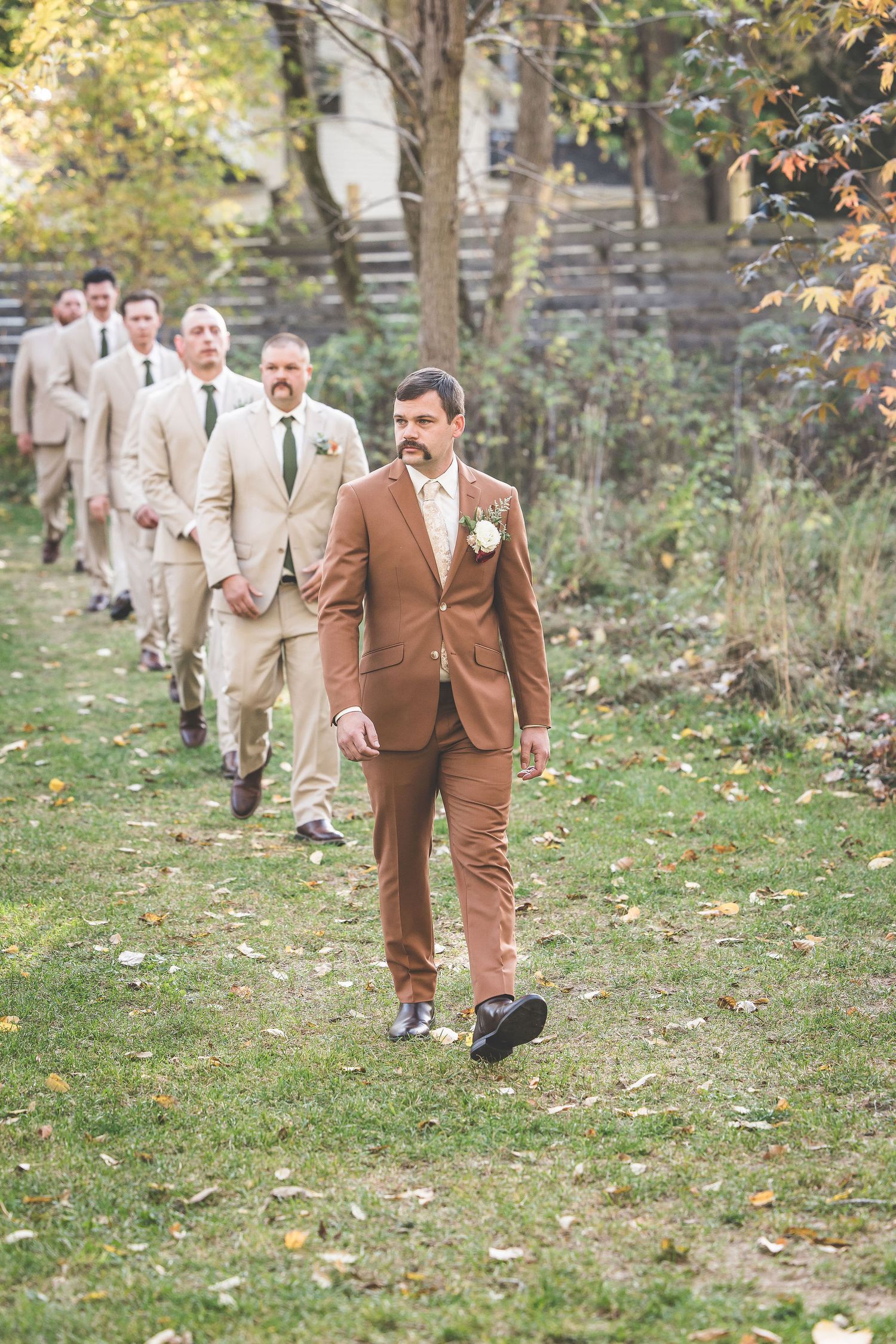 Groomsmen in beige suits follow a man in a brown suit walking down a garden path.