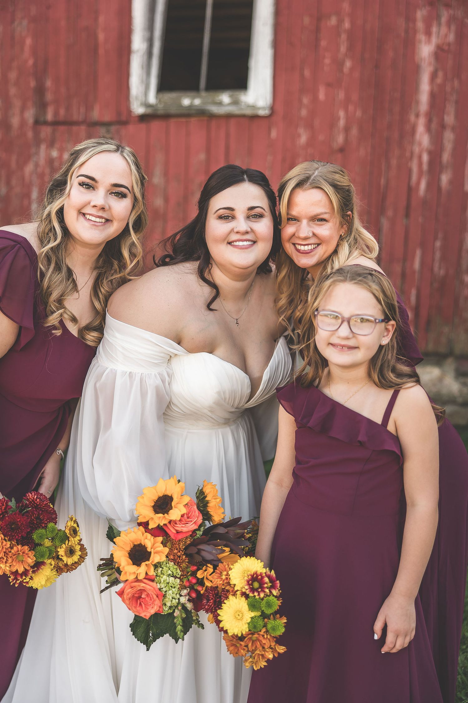 Wedding party members with sunflower bouquets pose together by red barn.