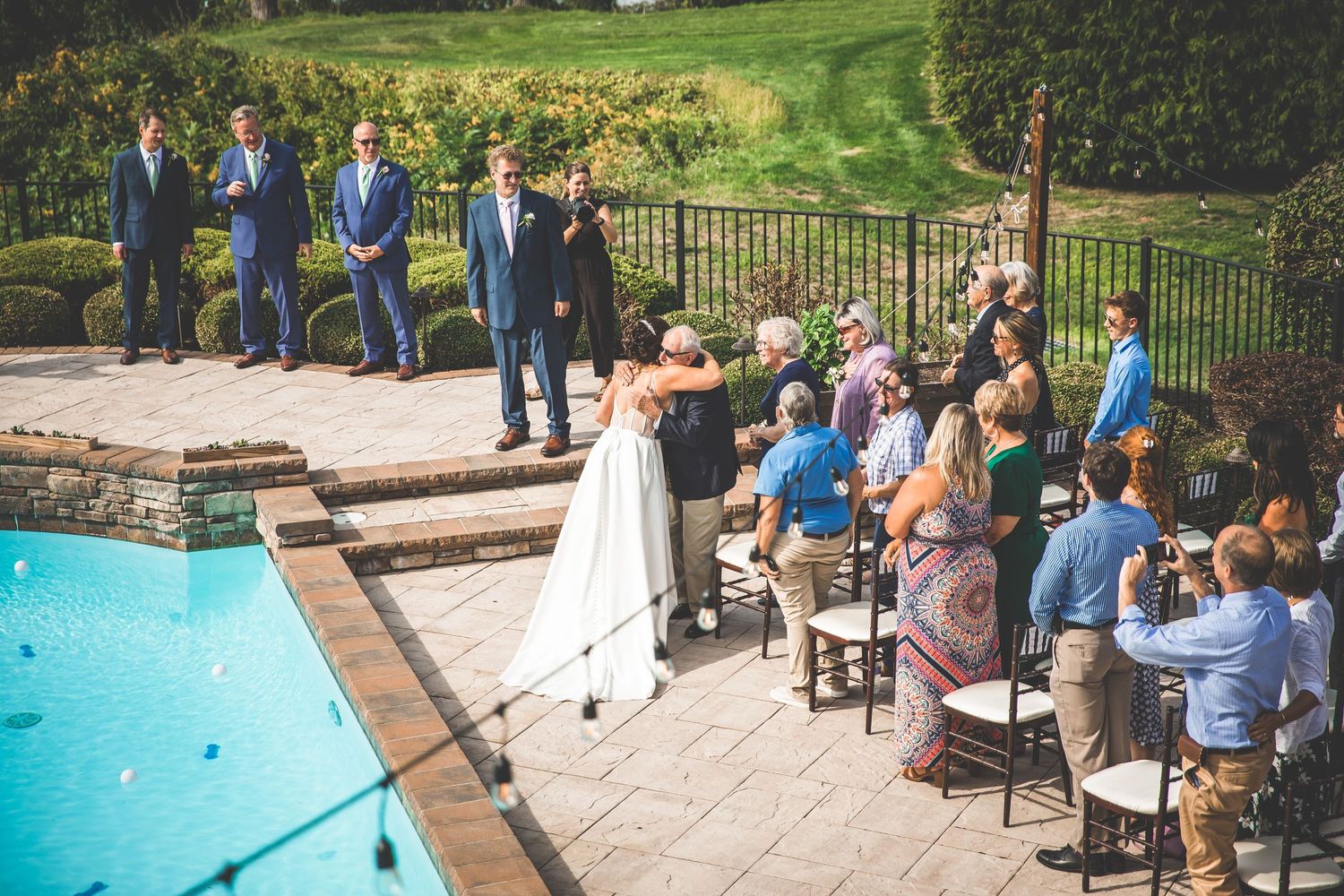 Outdoor wedding ceremony with guests seated around a swimming pool under blue skies.