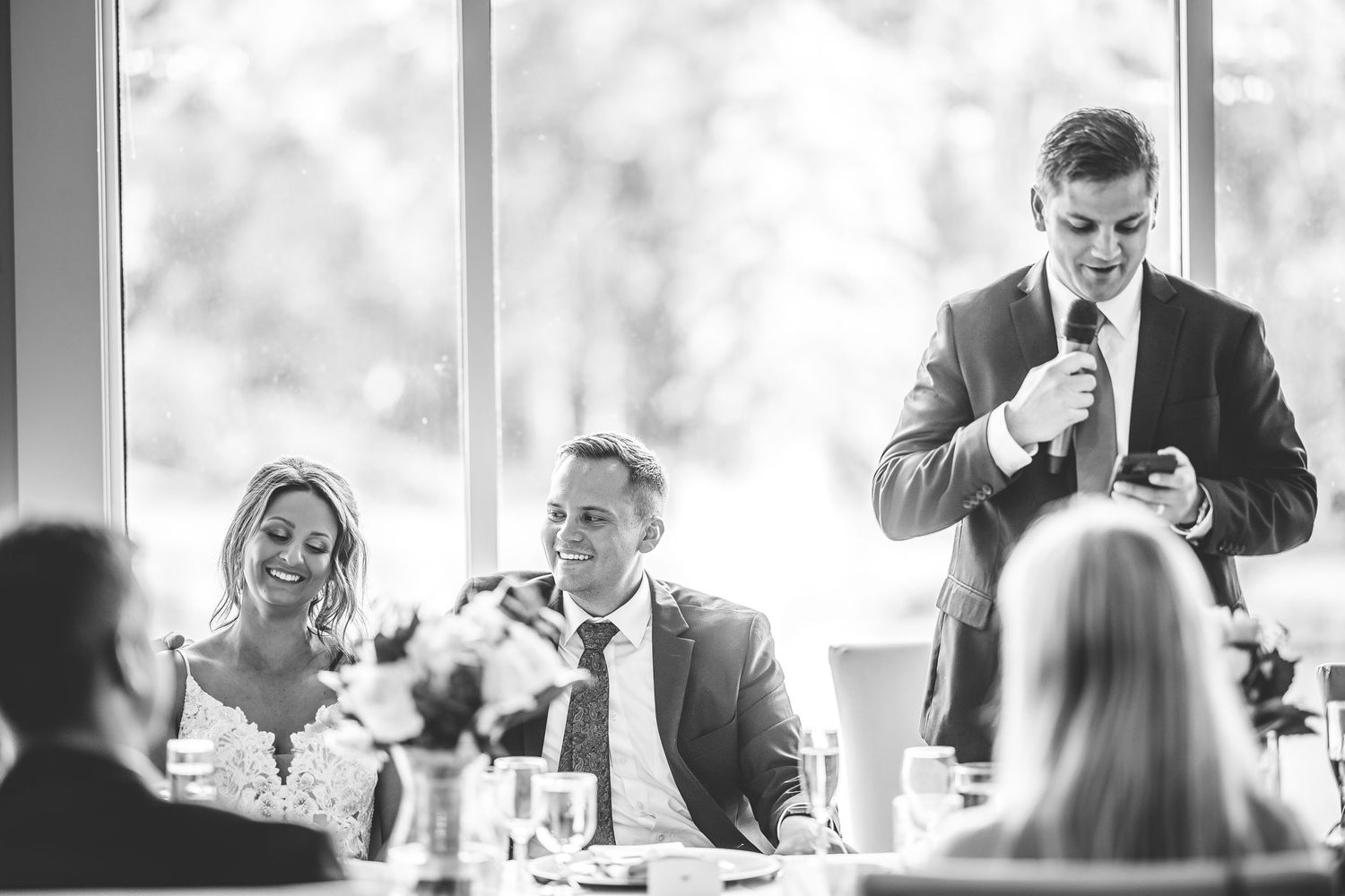 Reception guests enjoy dinner and conversation at elegantly set tables by large windows.