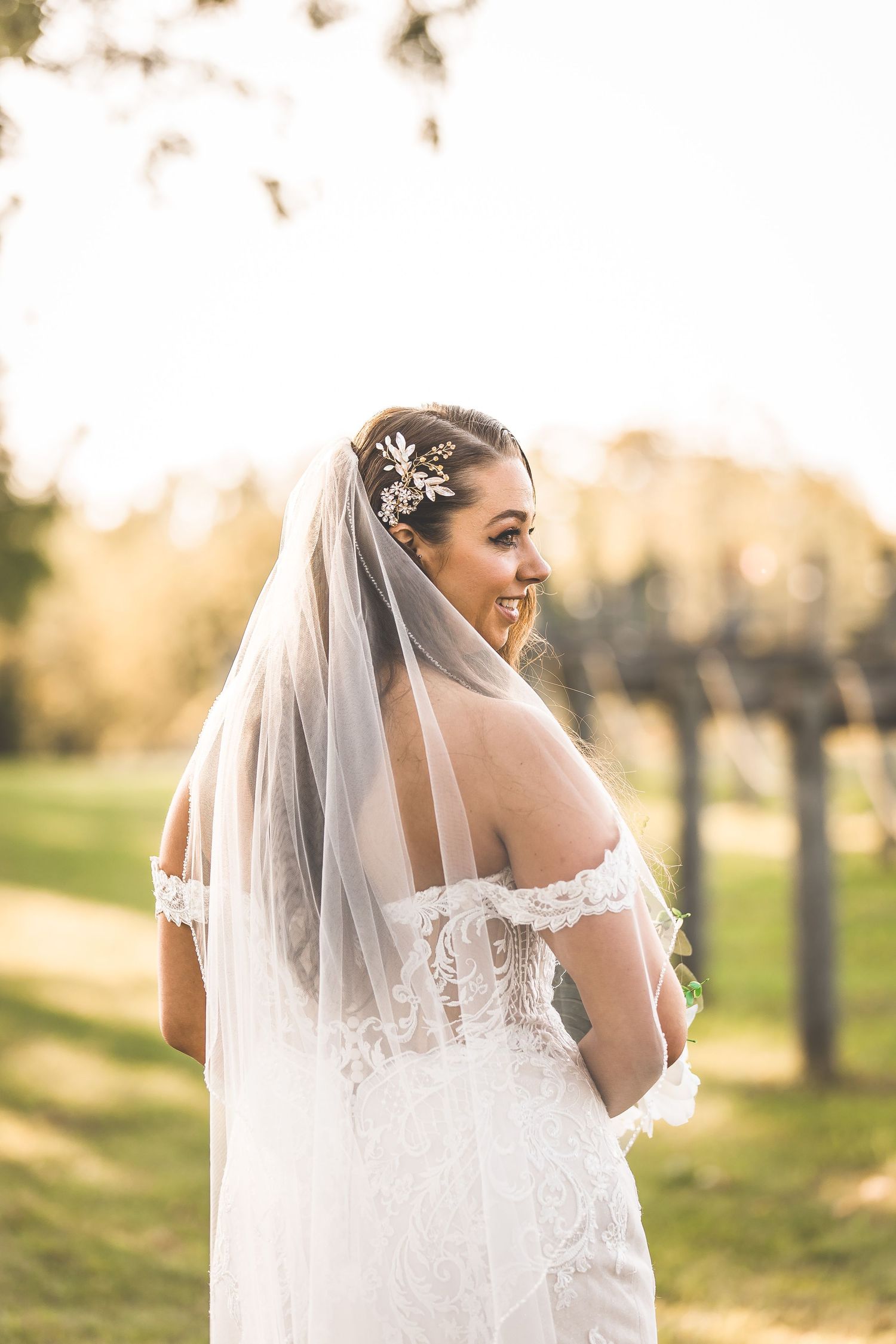 A bride in a lace wedding dress and veil poses in golden sunlight among trees.