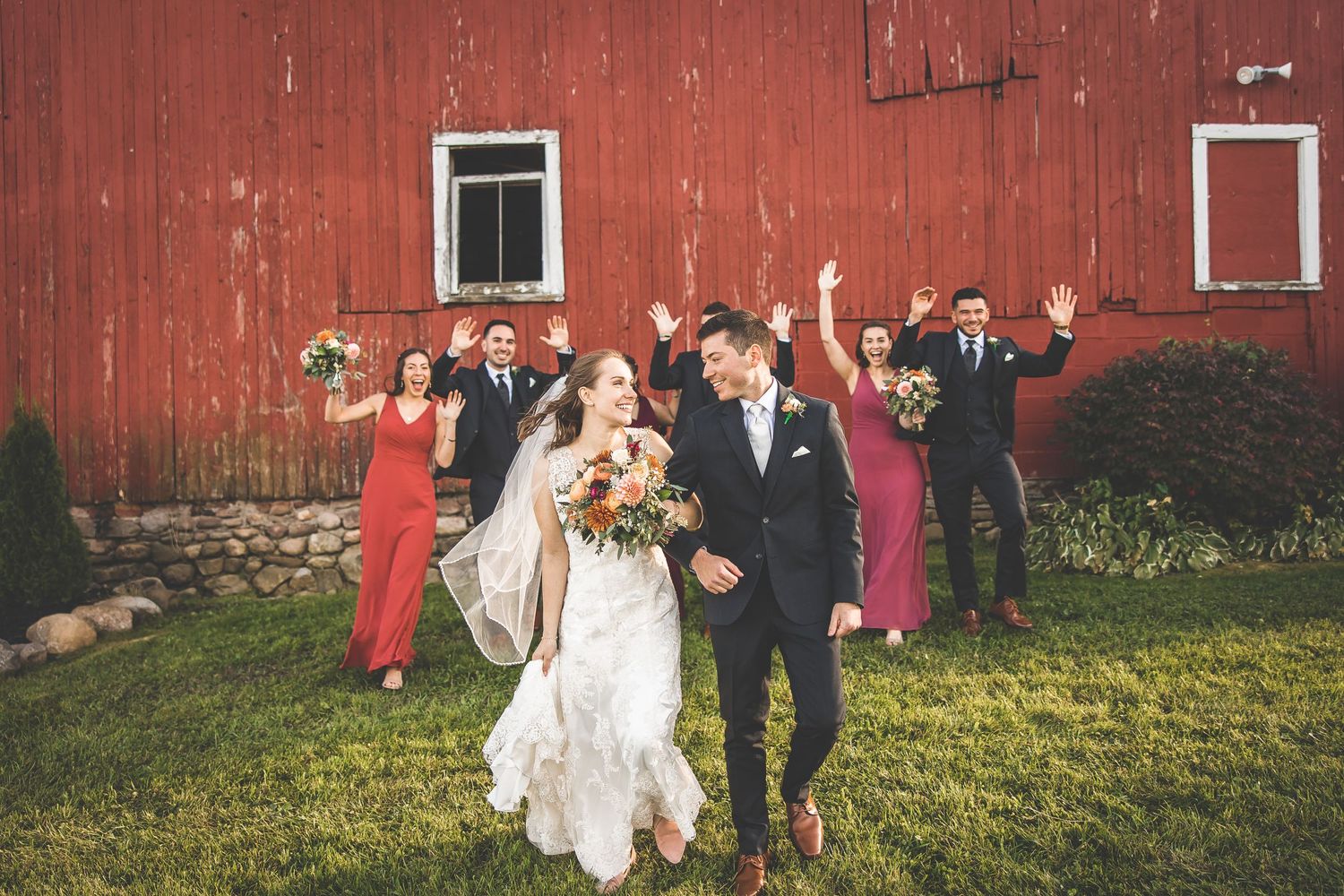 Wedding party poses in front of a rustic red barn on a grassy lawn.