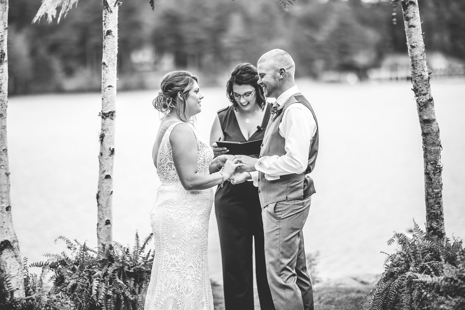 Black and white capture of wedding ceremony proceedings under rustic birch arch by lakeside.