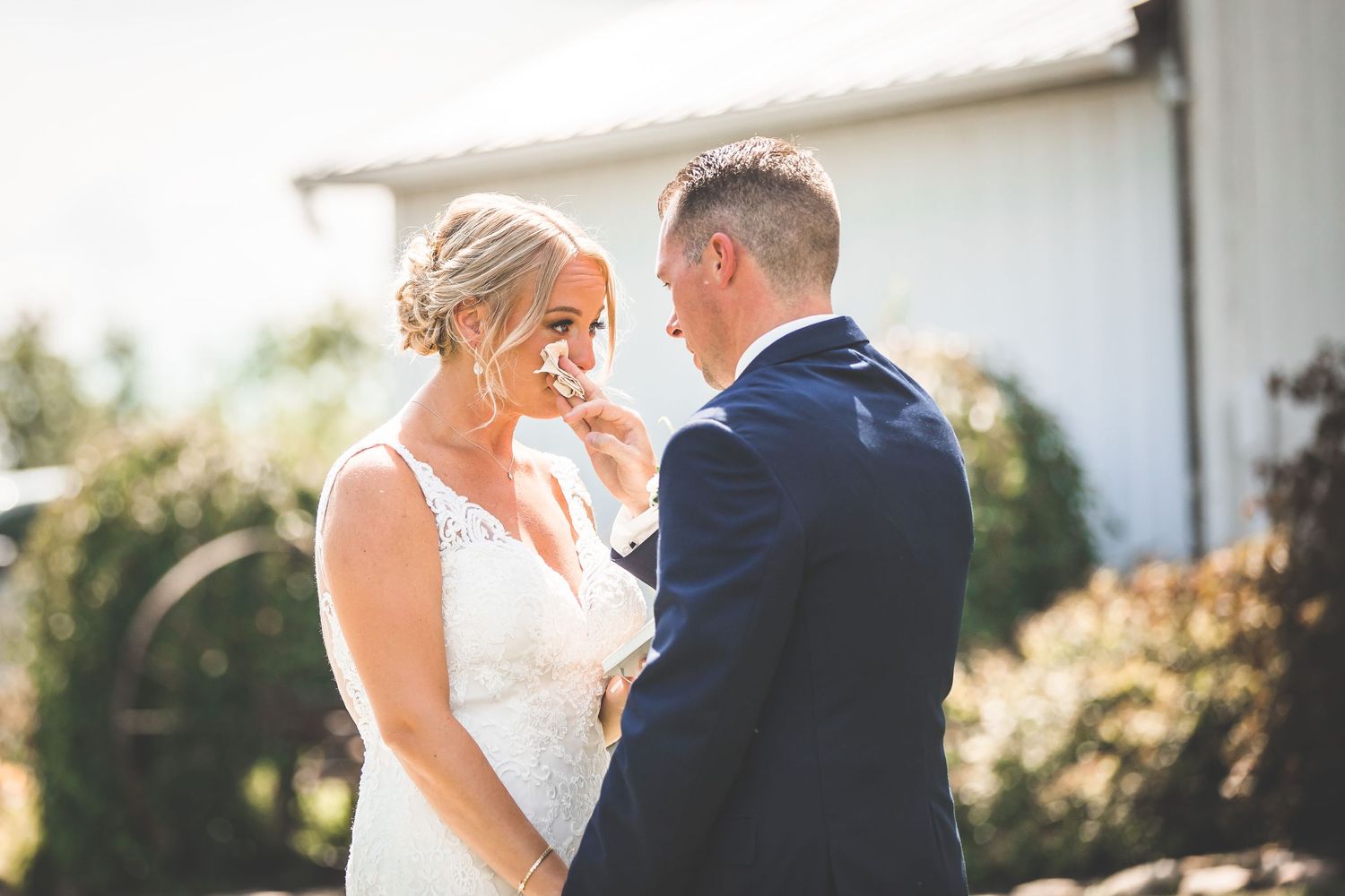 Sunlit outdoor wedding scene with a couple sharing an intimate moment near a white building.