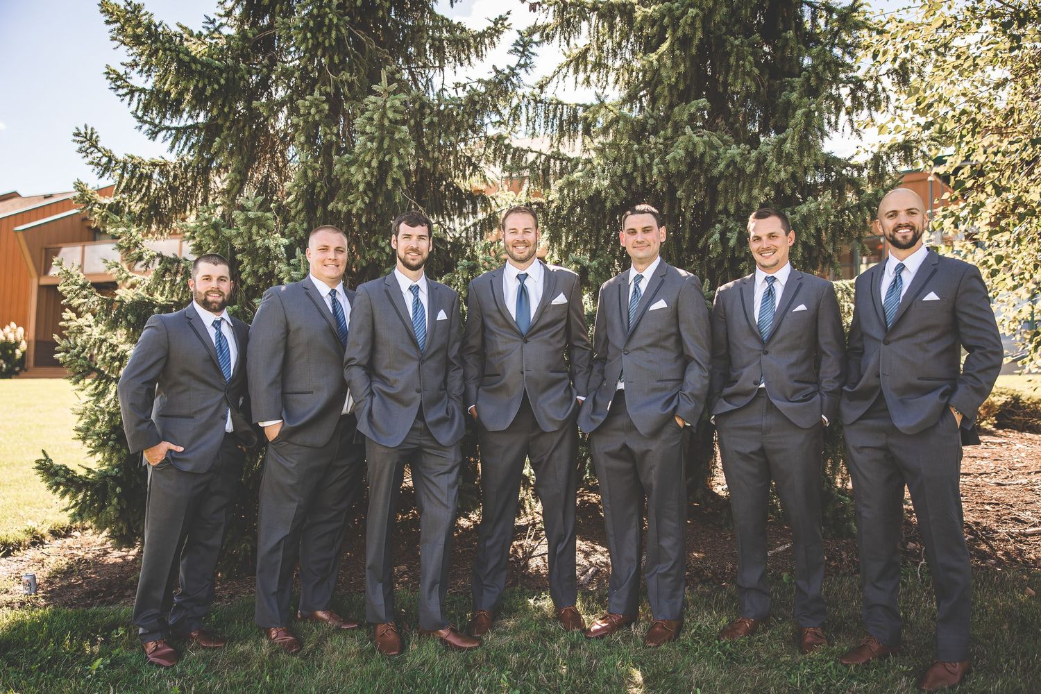 Large wedding party in matching gray suits and blue ties lined up outdoors by pine trees.