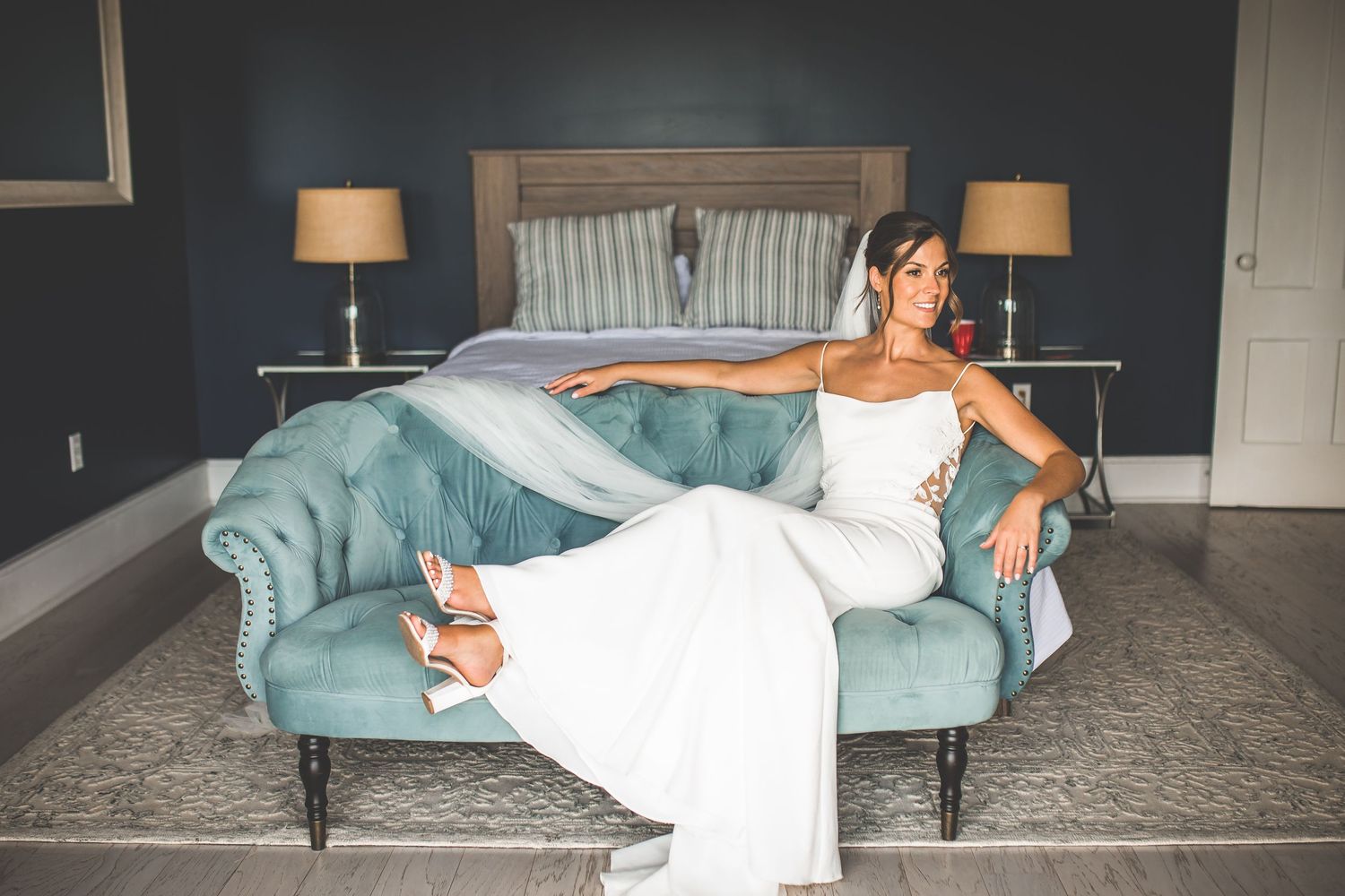 Bride relaxes on teal velvet couch in elegant room with dark walls.