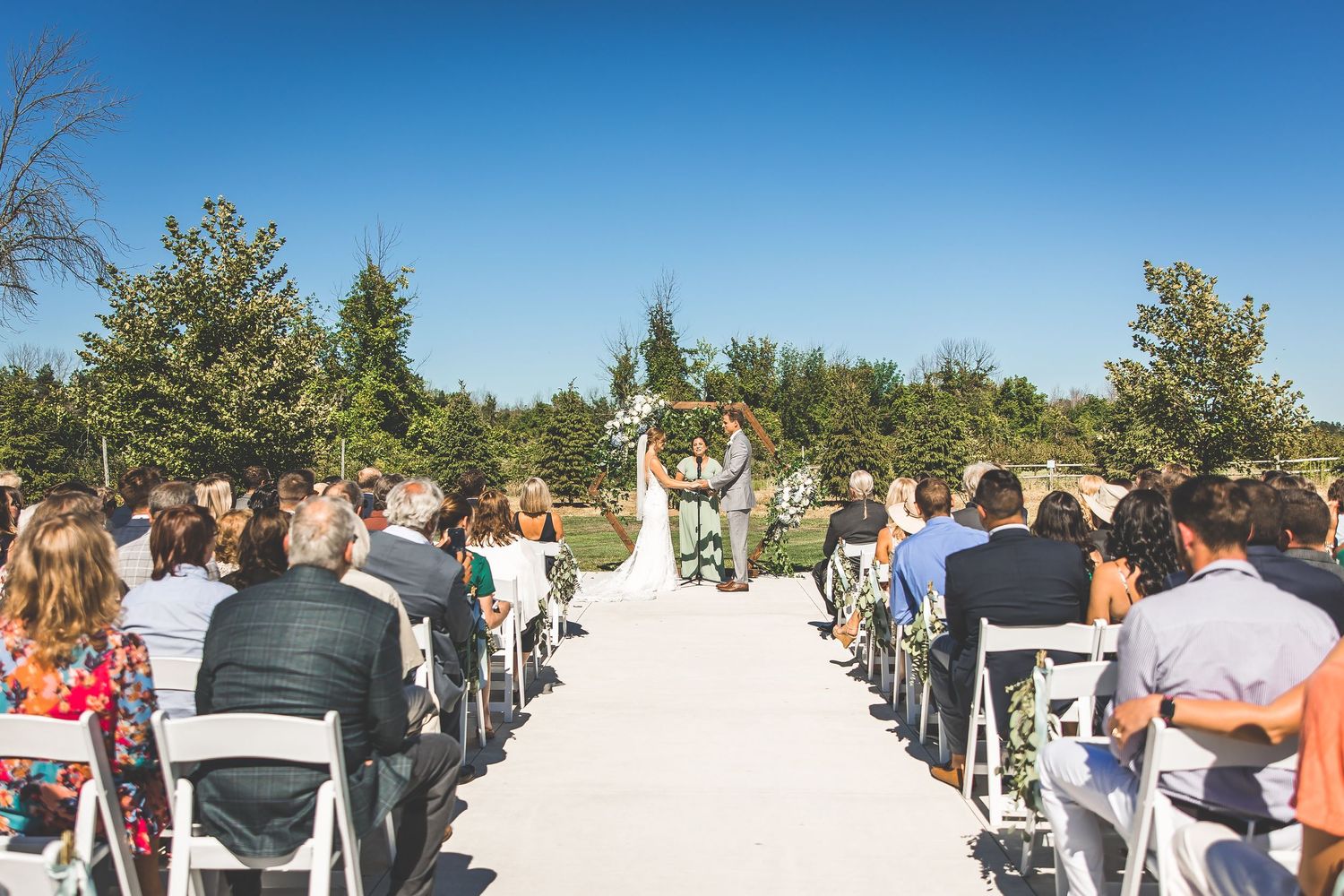 Wide view of outdoor wedding ceremony with white chairs and floral arch against blue sky.