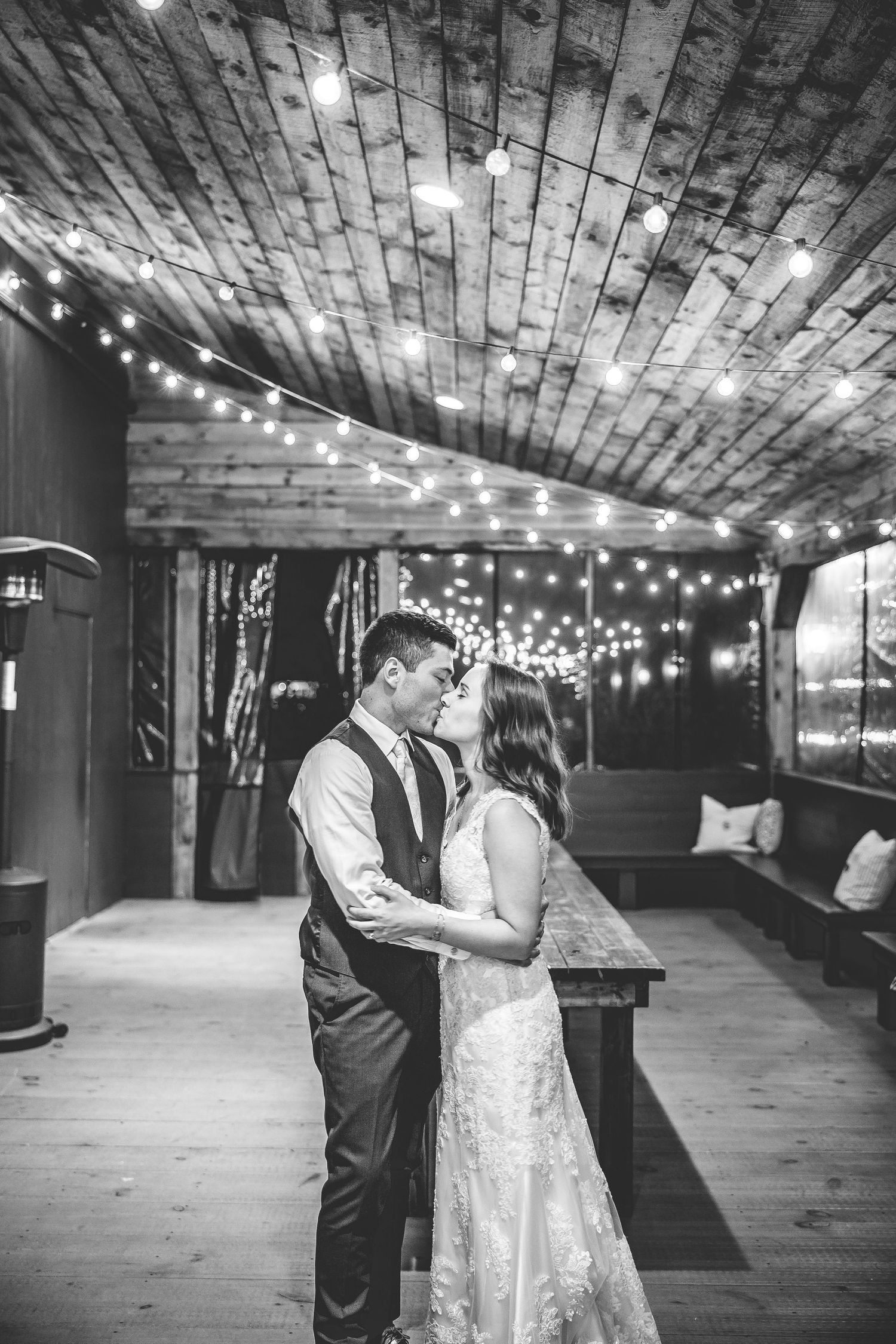Black and white photo of a couple dancing in a rustic venue with string lights overhead.