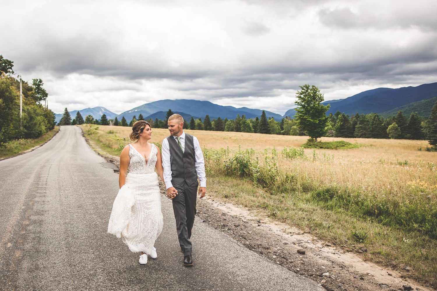 Wedding couple walks hand in hand down a rural mountain road surrounded by fields.