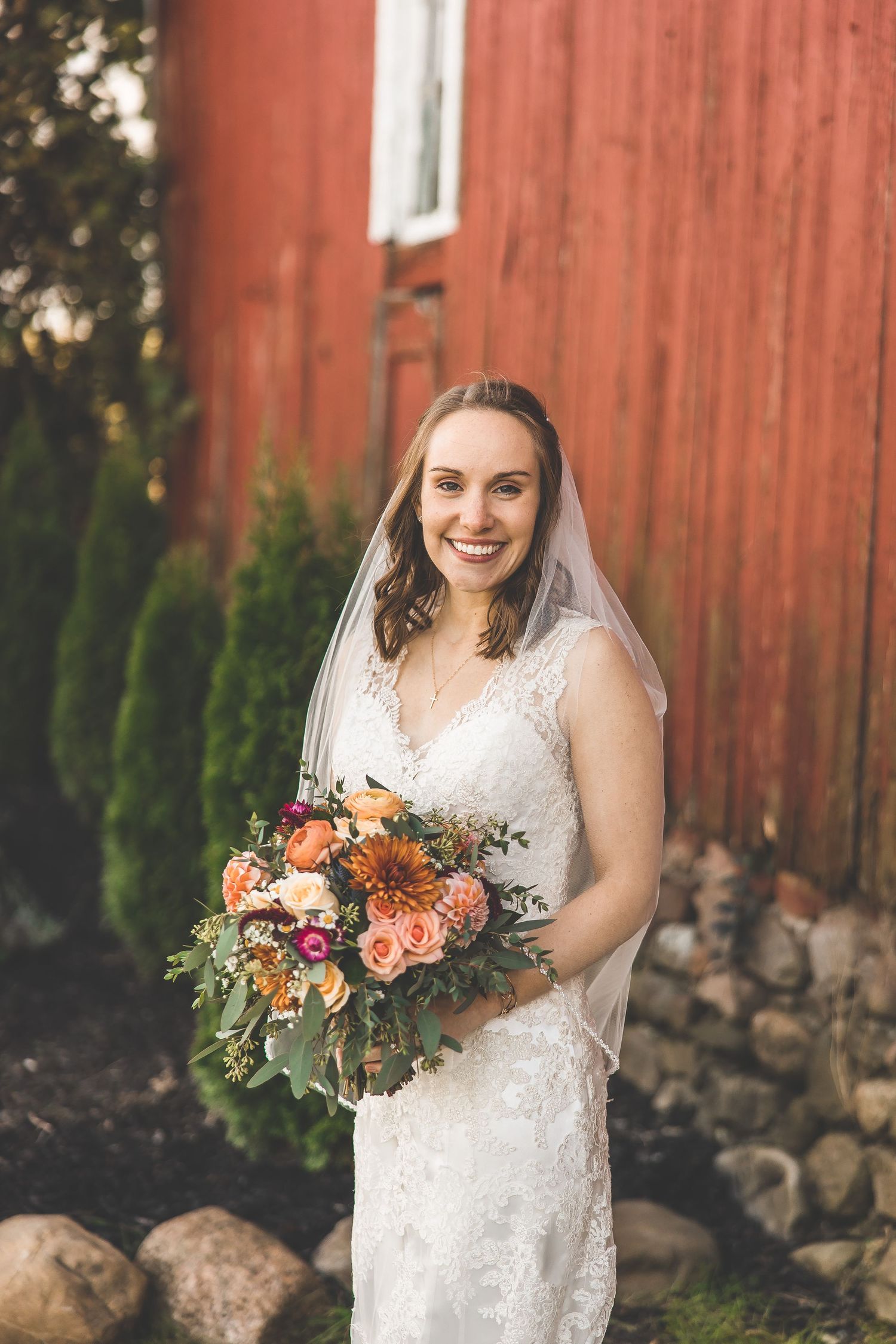 Bride in lace dress holding autumn-colored bouquet against red barn background.