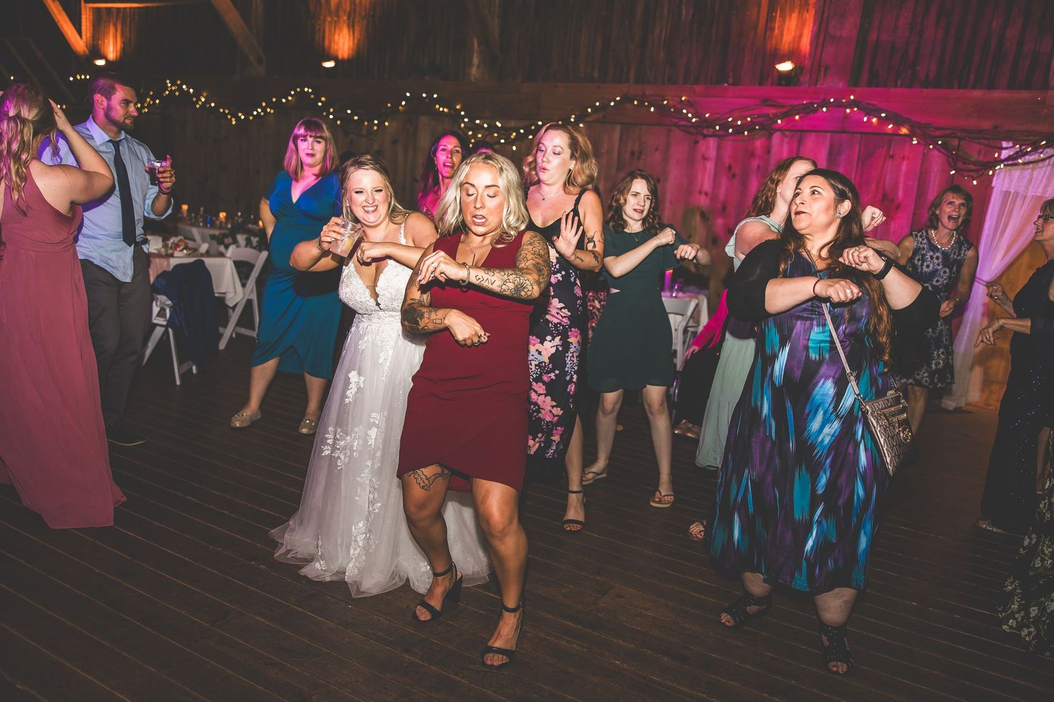 Guests dance energetically at a wedding reception with pink decorative lighting and string lights.